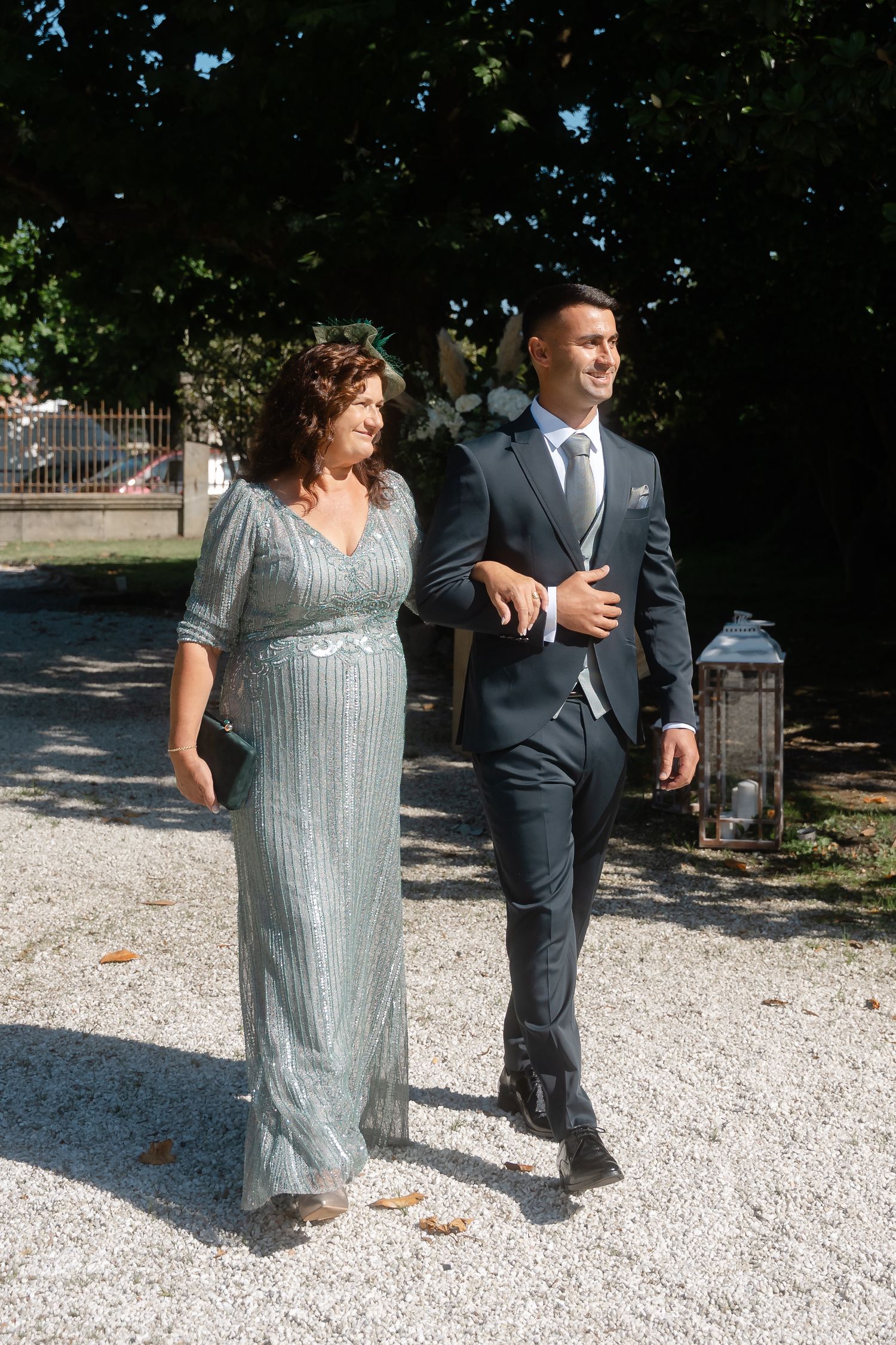 A woman in a sparkly sage gown walks arm in arm with a man wearing a navy suit on a gravel path outdoors.