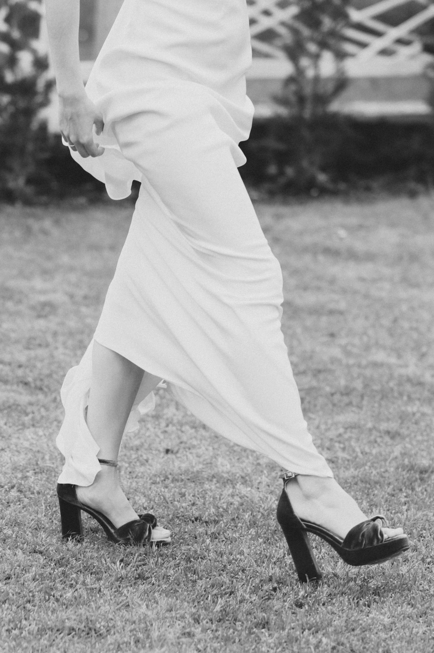 A black and white photo showing the lower half of a wedding dress and black heels walking on grass.