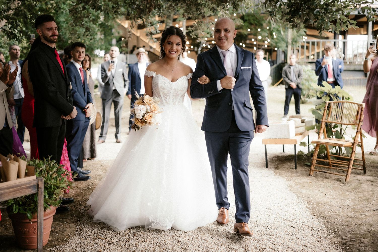 A wedding ceremony outdoors with guests looking on as a couple walks down a path lined with rustic decor.