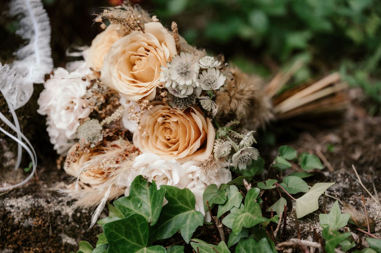 A delicate dried wedding bouquet with peach roses and dried flowers rests on soil beside green ivy leaves.