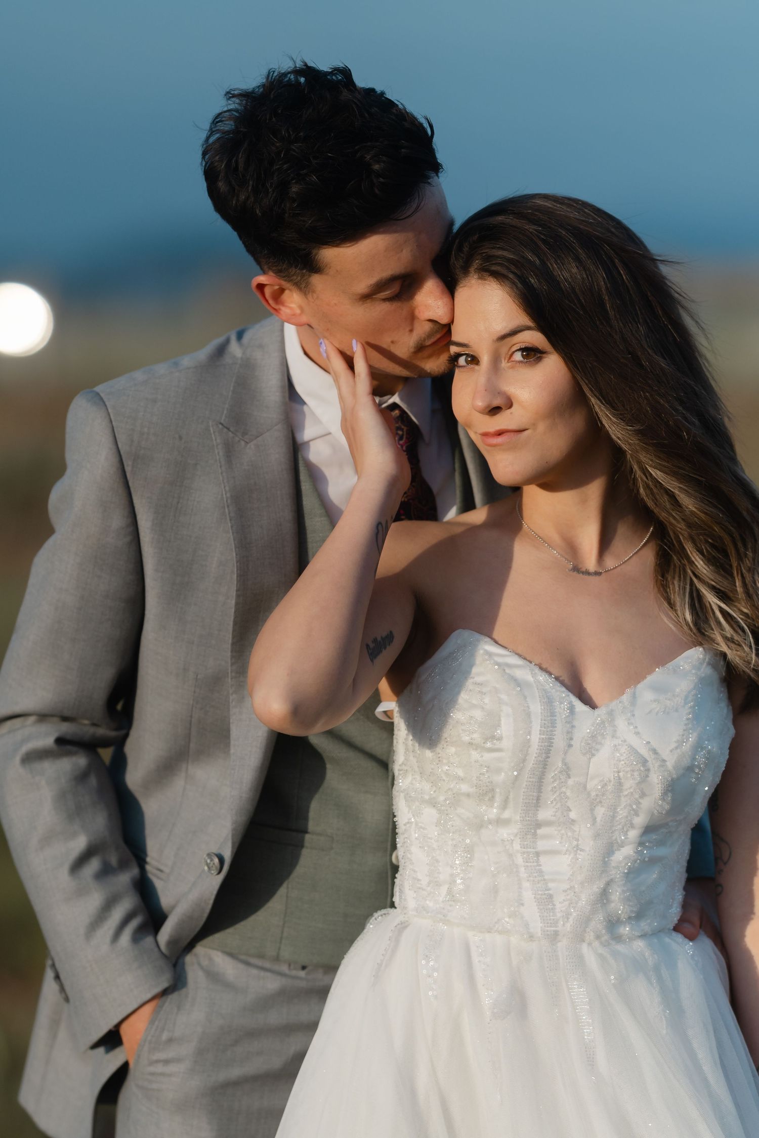 Groom in gray suit tenderly kissing bride's cheek as she gazes forward in a white strapless wedding gown.