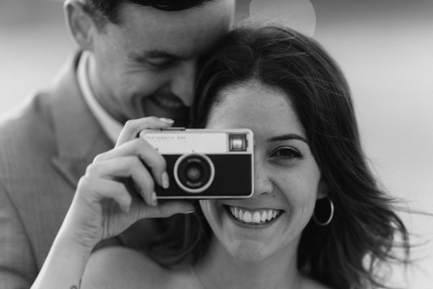 Black and white photo of a smiling woman holding a vintage camera while a man stands behind her.