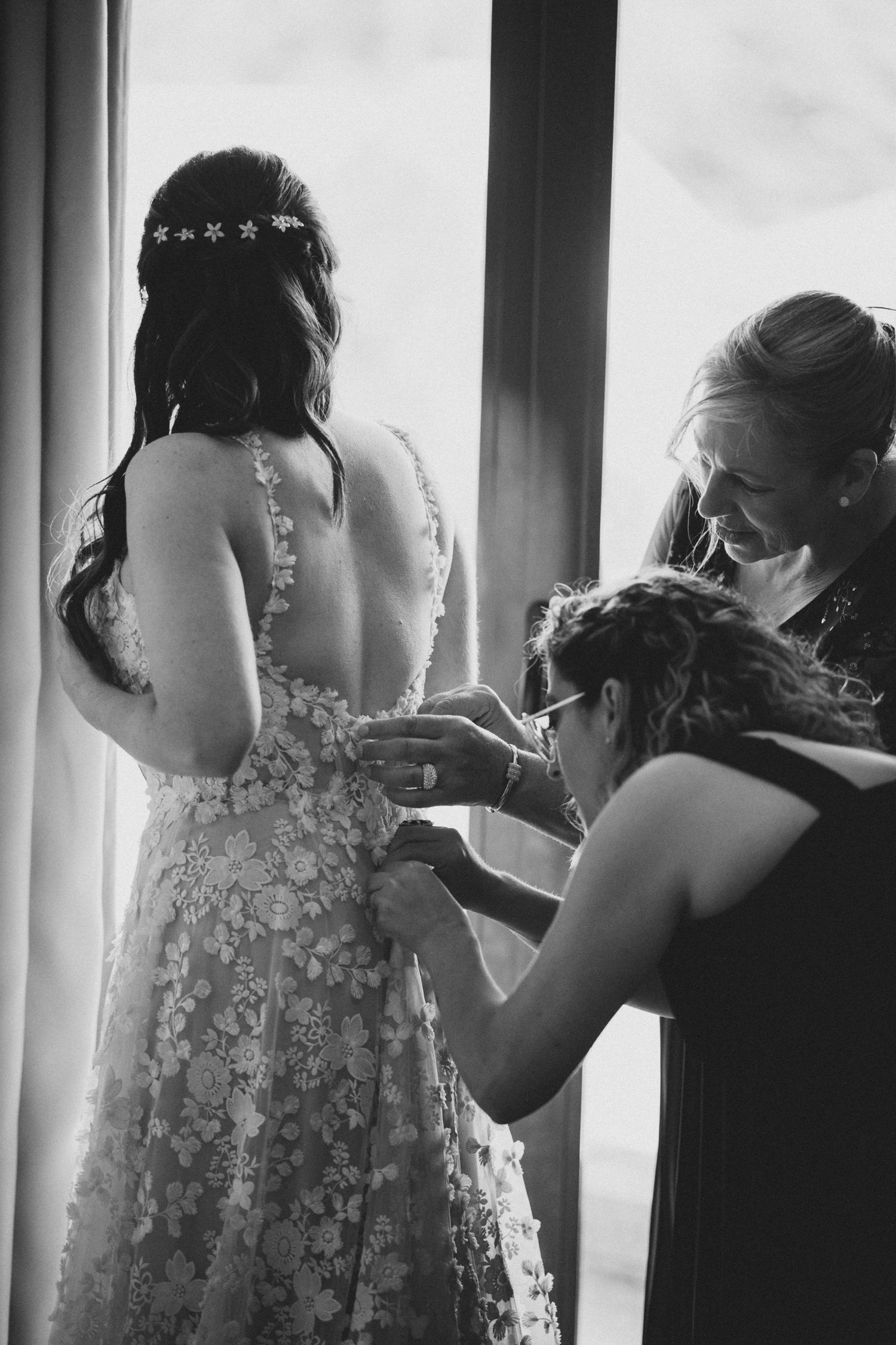 A black and white photo showing final dress preparations by a window, with delicate floral details on the gown.