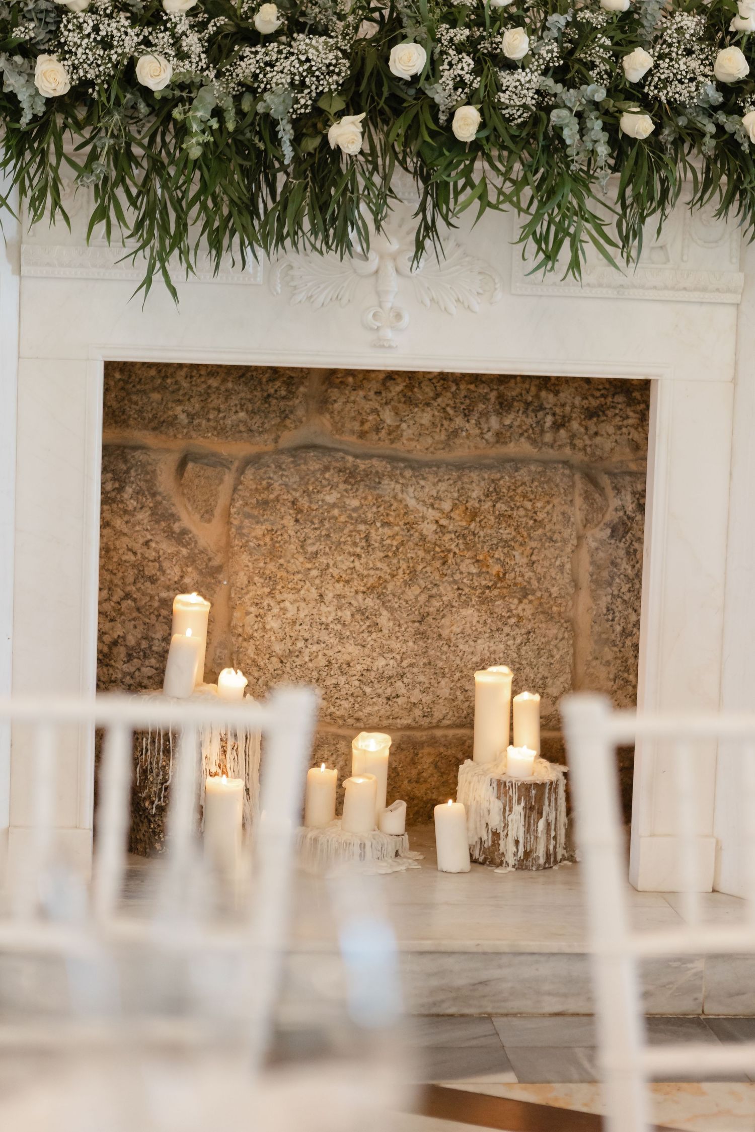 White pillar candles line a stone wall with lush greenery and floral decorations hanging above in an elegant venue setting.