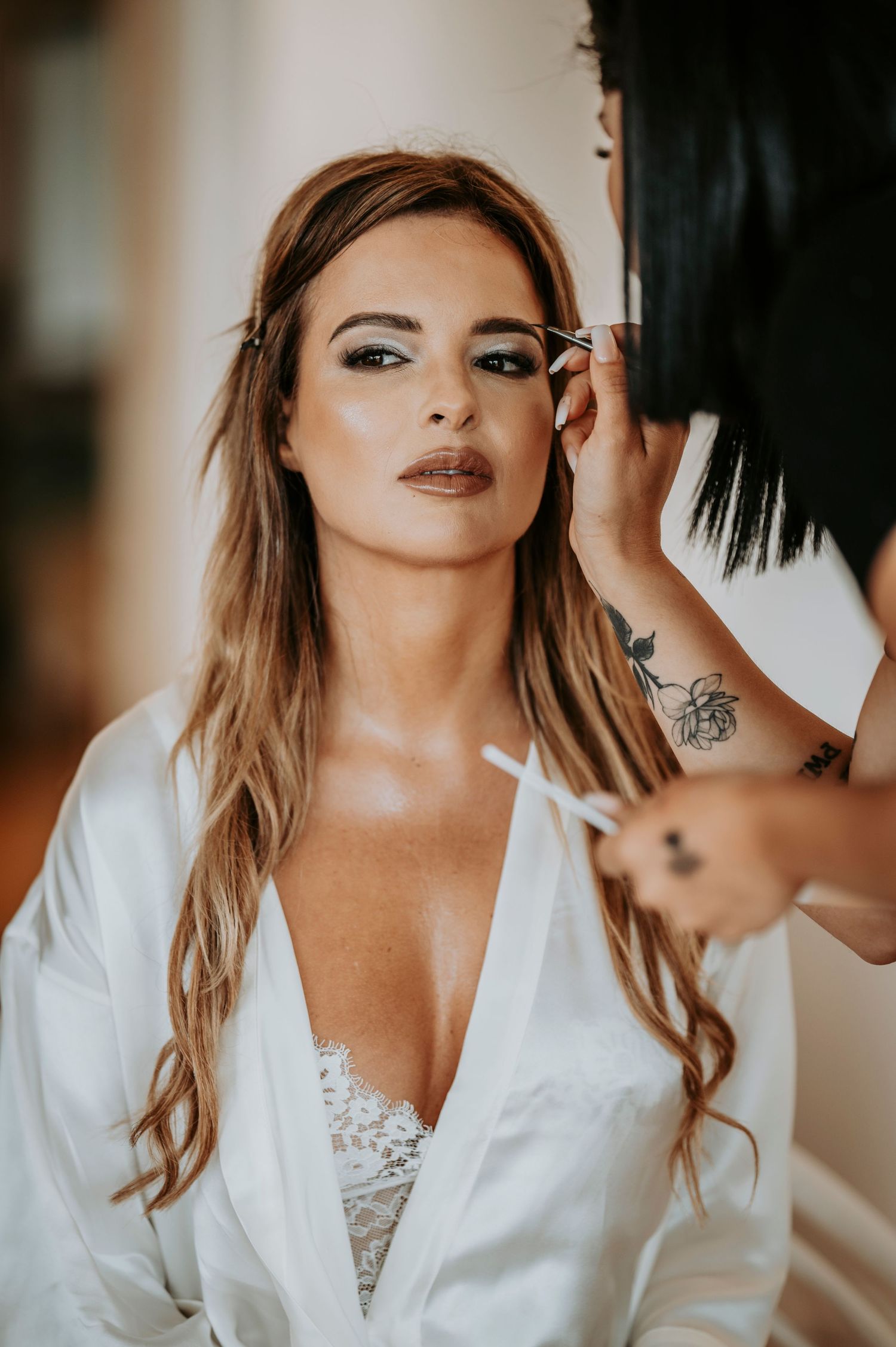 A makeup artist applies finishing touches to a model wearing a white robe and lace top during a beauty session.