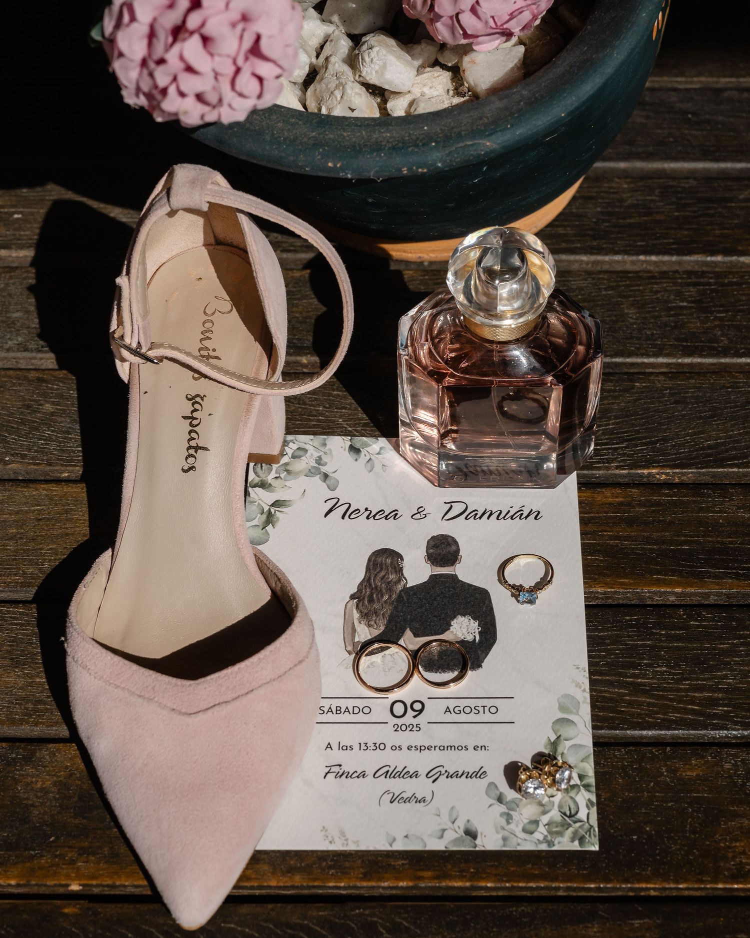 Wedding details featuring a pink high heel shoe, perfume bottle, and invitation card on a wooden surface.