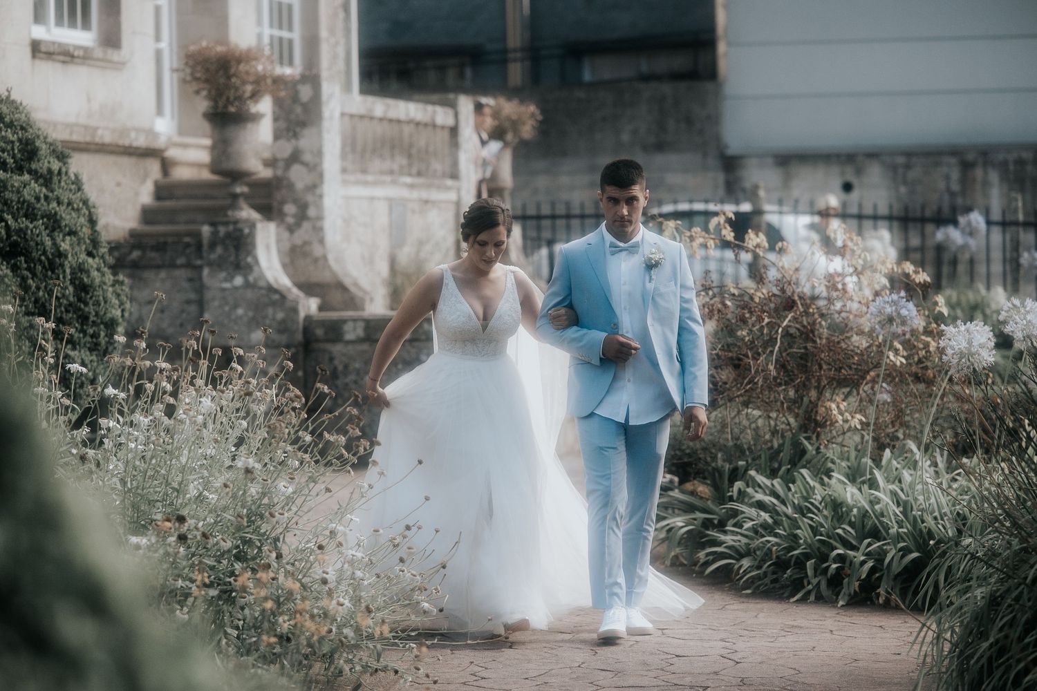 A couple in wedding attire walks through a lush garden setting with soft natural lighting.