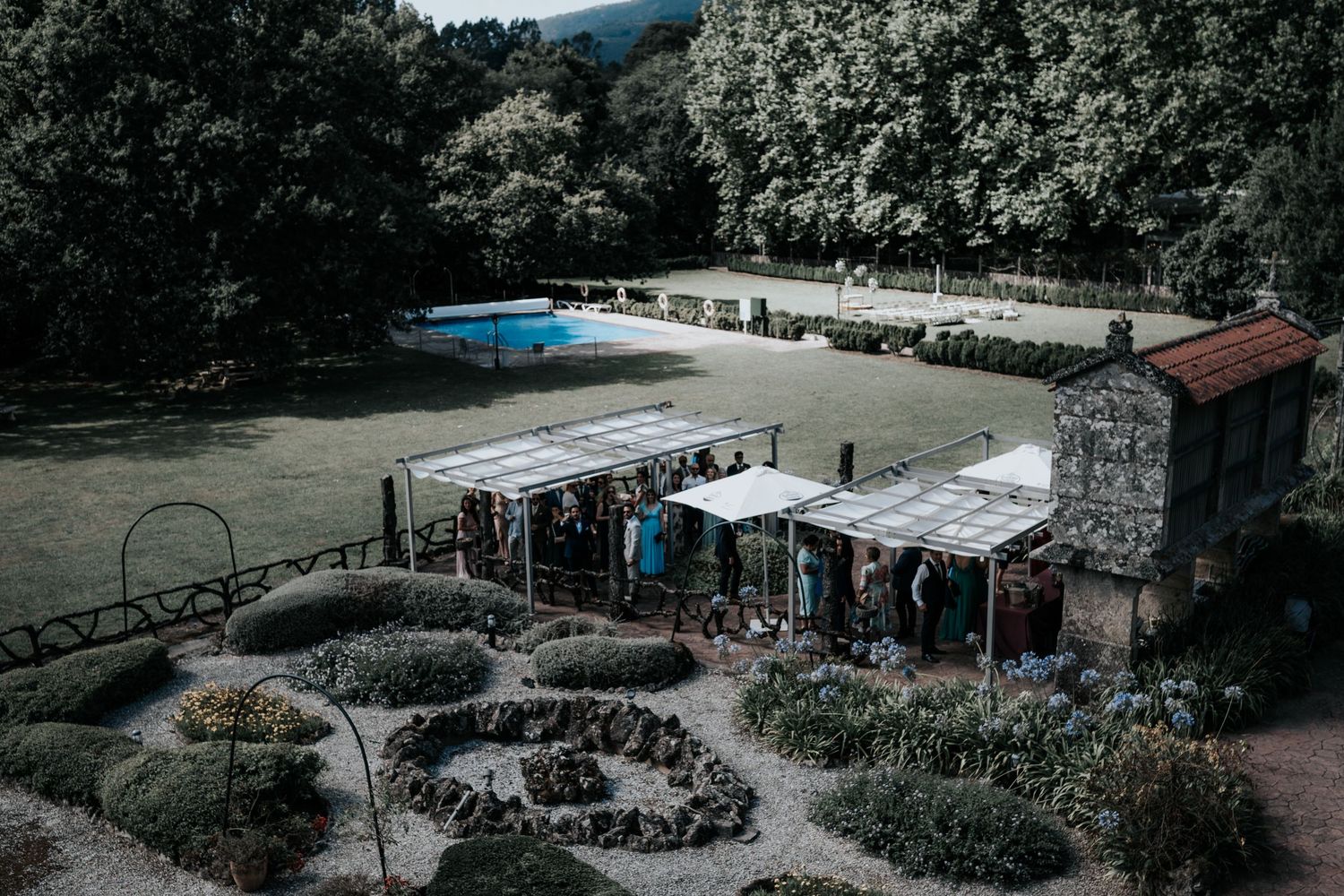 Aerial view of an outdoor venue with a pool, garden paths and vendor tents surrounded by lush forest.