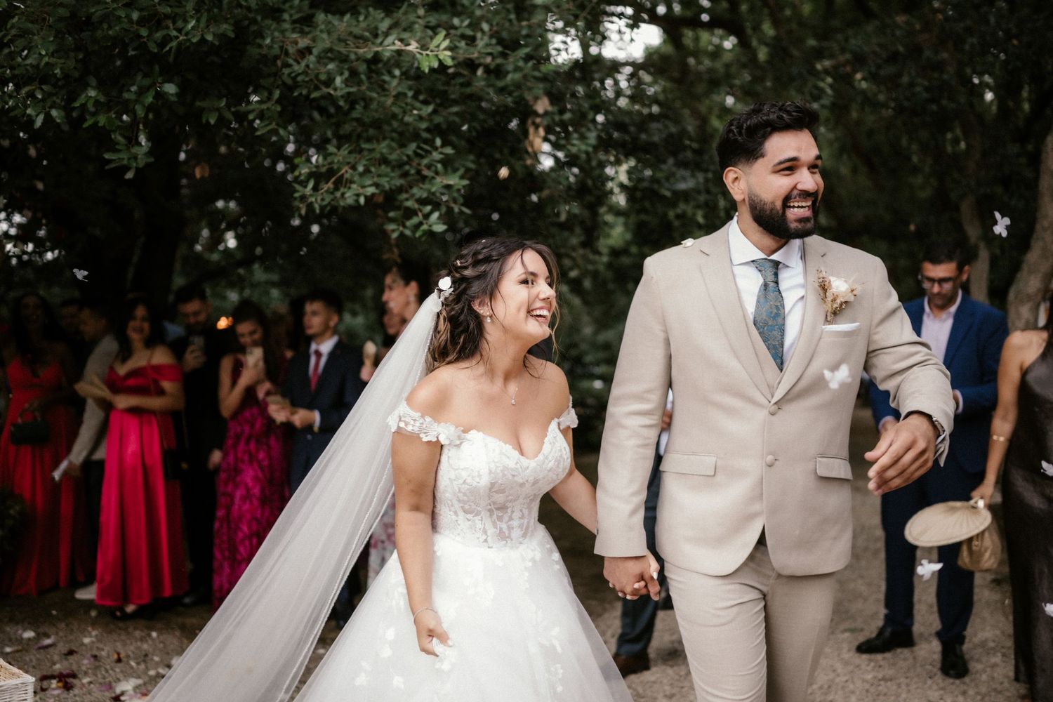A joyful wedding couple walking together outdoors with guests in colorful attire in the background.