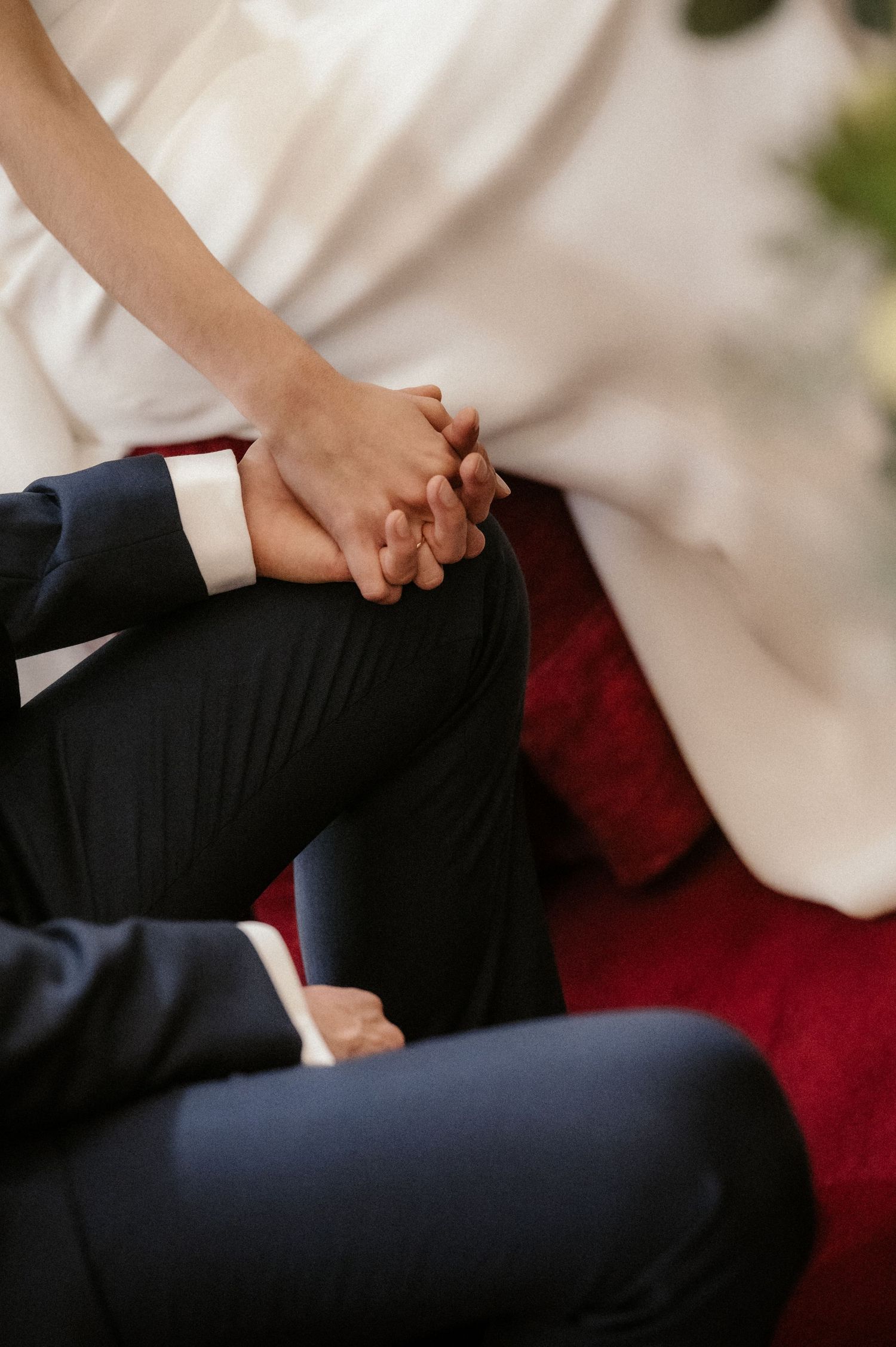 Close-up of a couple holding hands while sitting on a red velvet couch at their wedding.