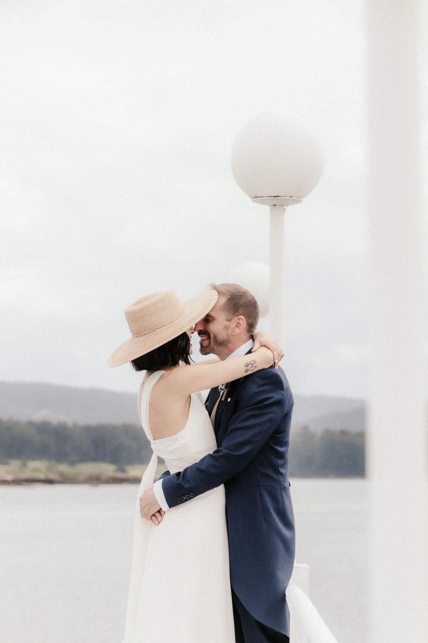 A couple shares a tender moment by the waterfront, with the bride wearing an elegant white gown.