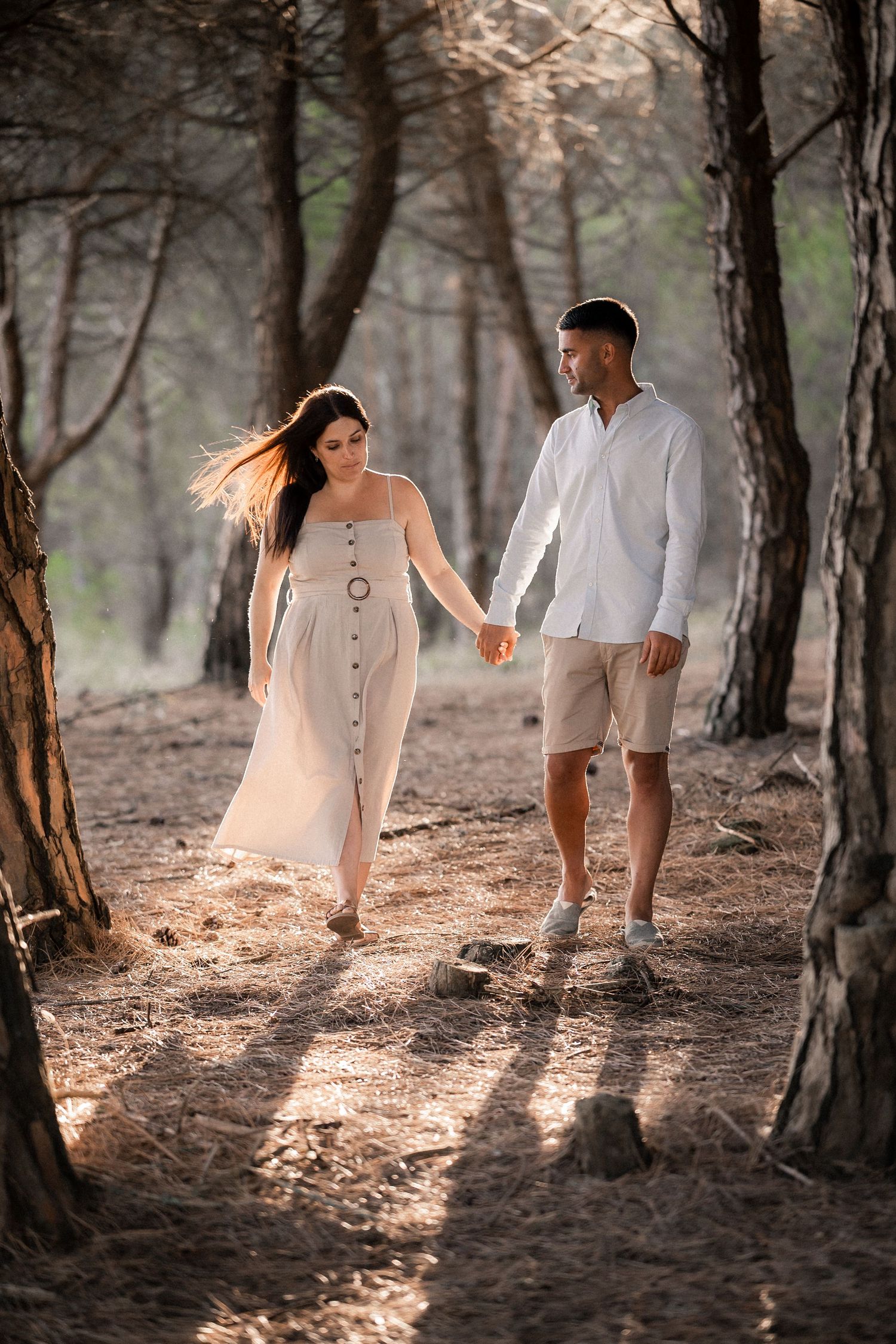 Romantic couple walking hand in hand through sunlit forest path in light summer attire.
