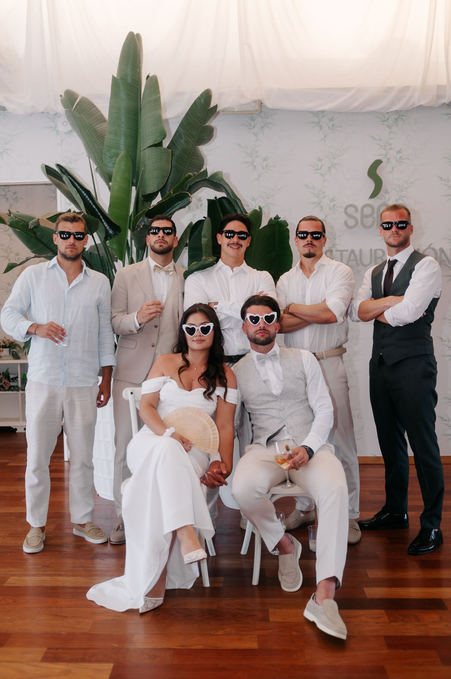 Wedding party members in white attire strike fun poses on wooden dance floor at reception.