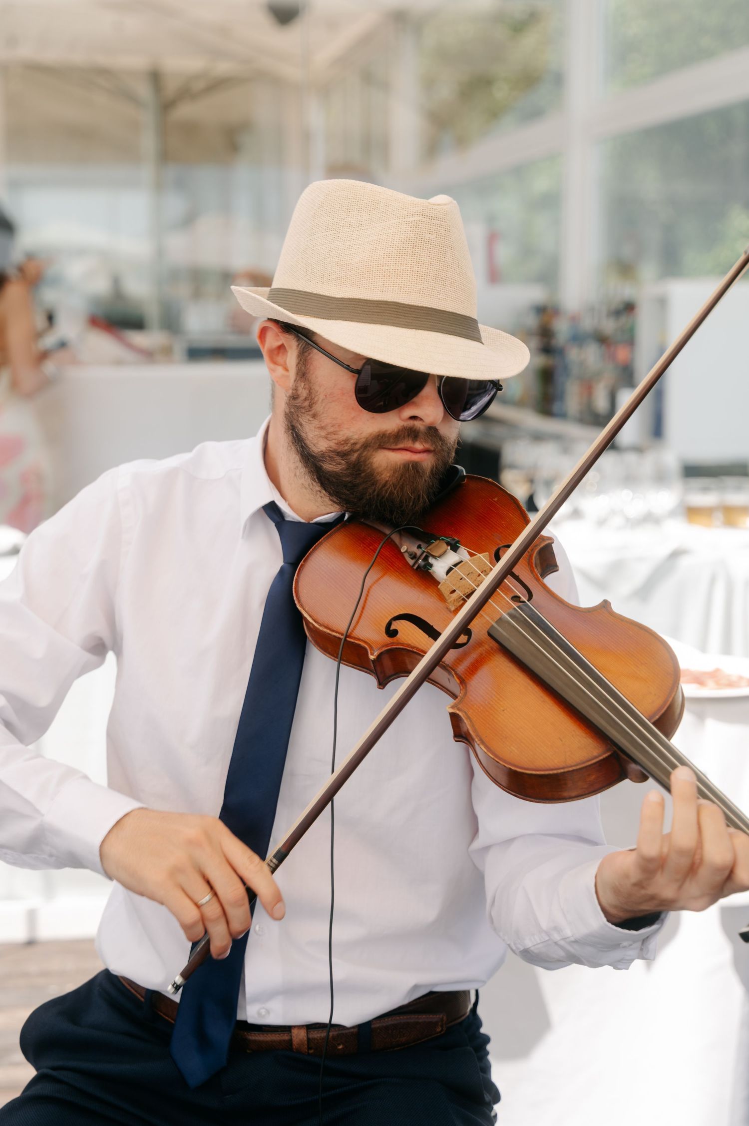A violinist in a white shirt and blue tie performs while wearing sunglasses and a light-colored hat.