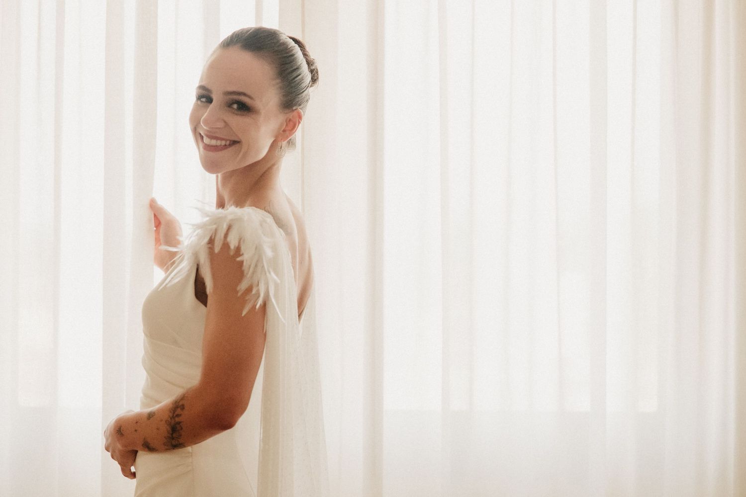 A bride in a white wedding gown poses elegantly against a light curtain background.