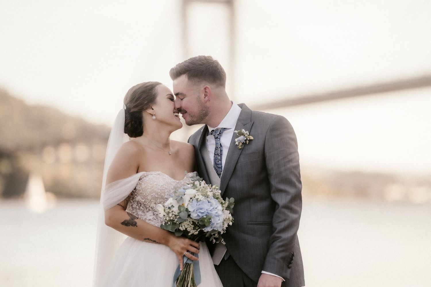 A romantic wedding couple shares an intimate moment in front of a blurred bridge background.