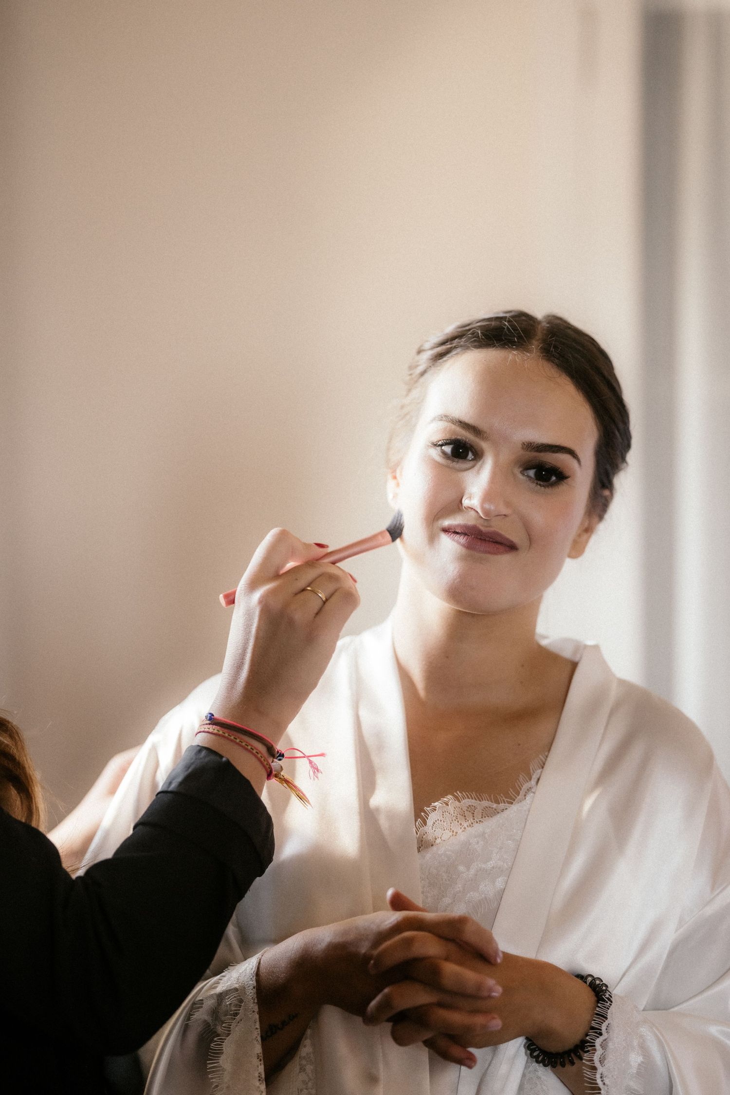 A makeup artist applies finishing touches to a bride wearing a white silk robe on her wedding day.