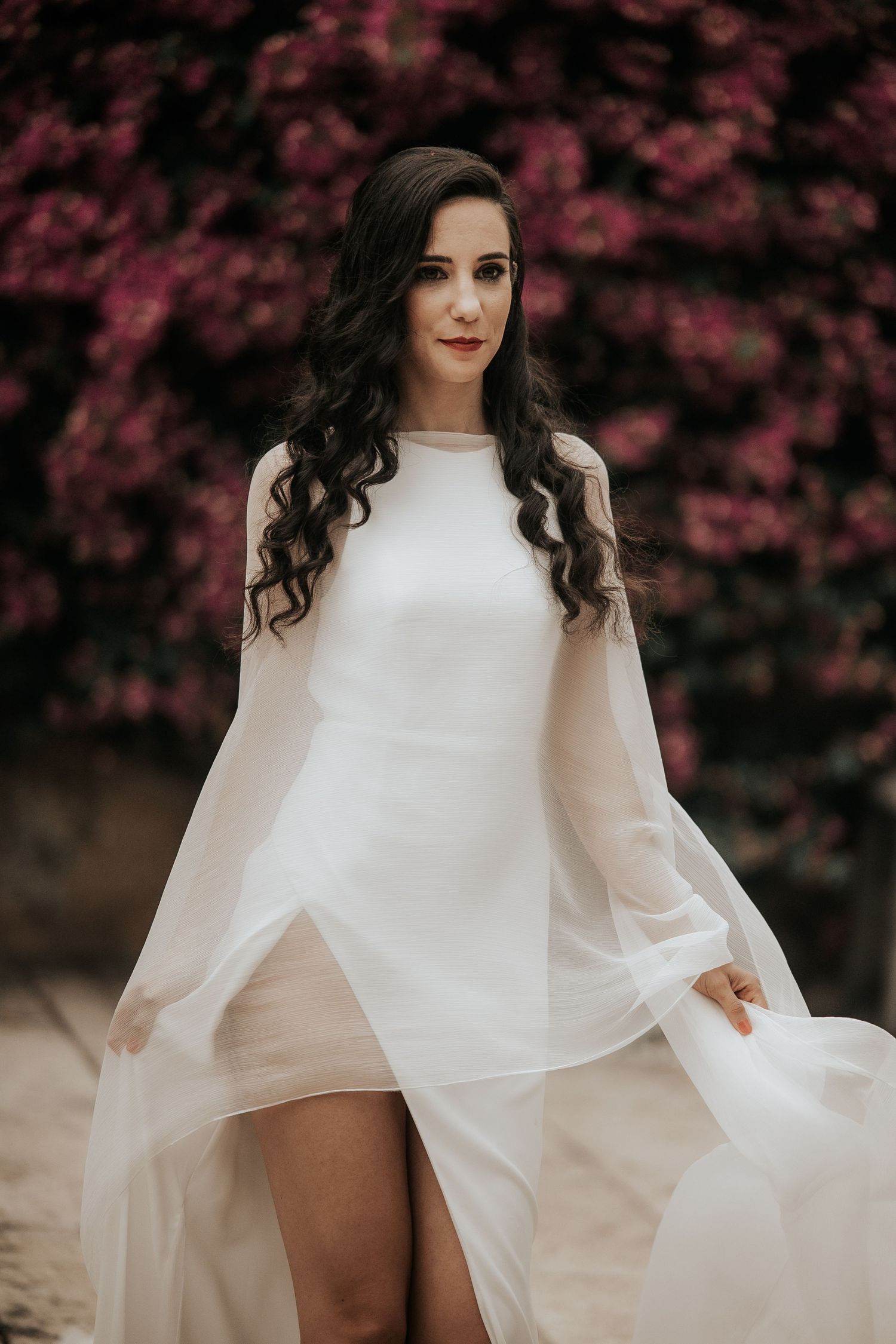Elegant white bridal cape dress photographed against pink flowering trees in romantic outdoor setting.