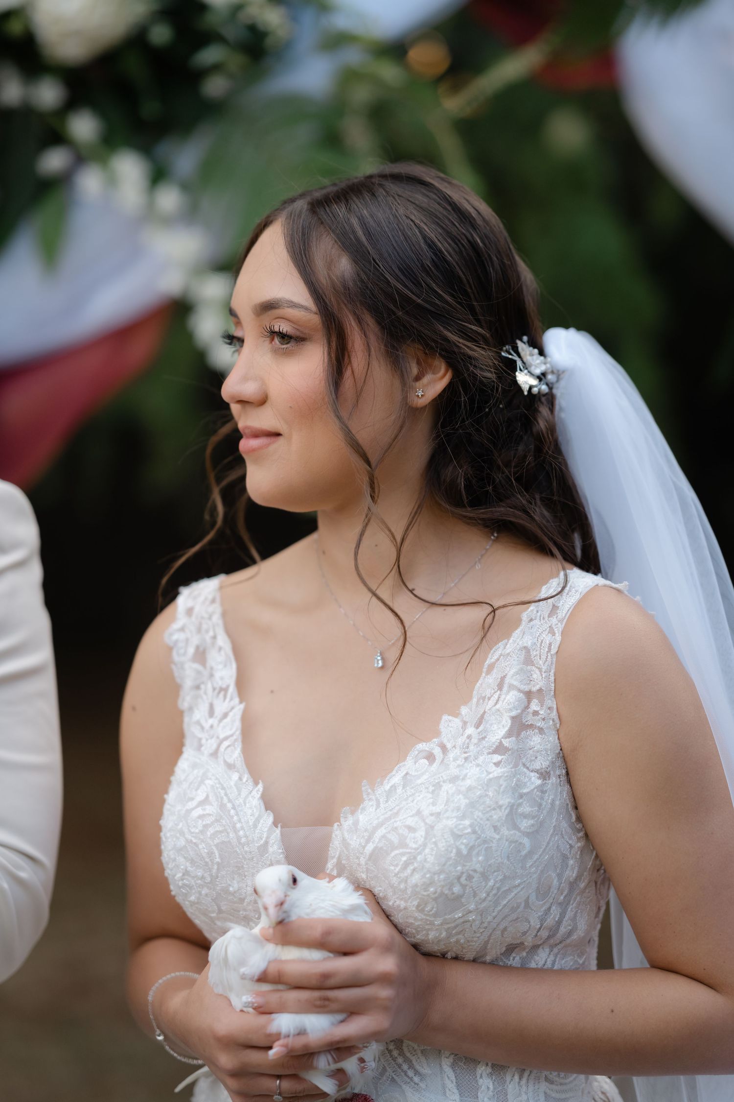 A bride in a white lace wedding dress holds a white dove in an outdoor garden setting.