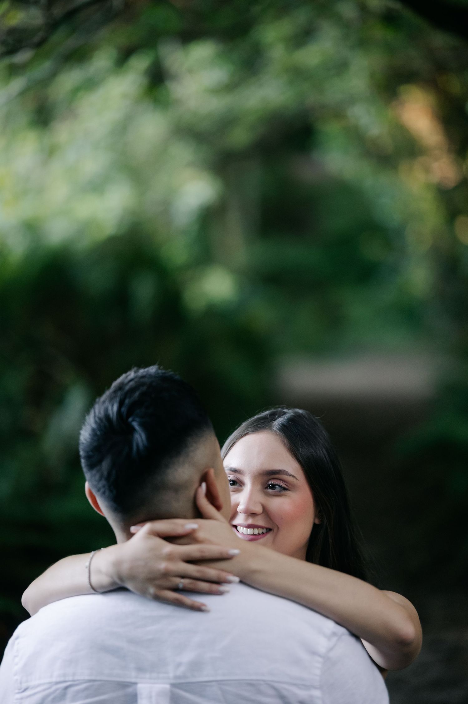 A romantic couple embraces outdoors in a garden setting with soft natural lighting and greenery in the background.