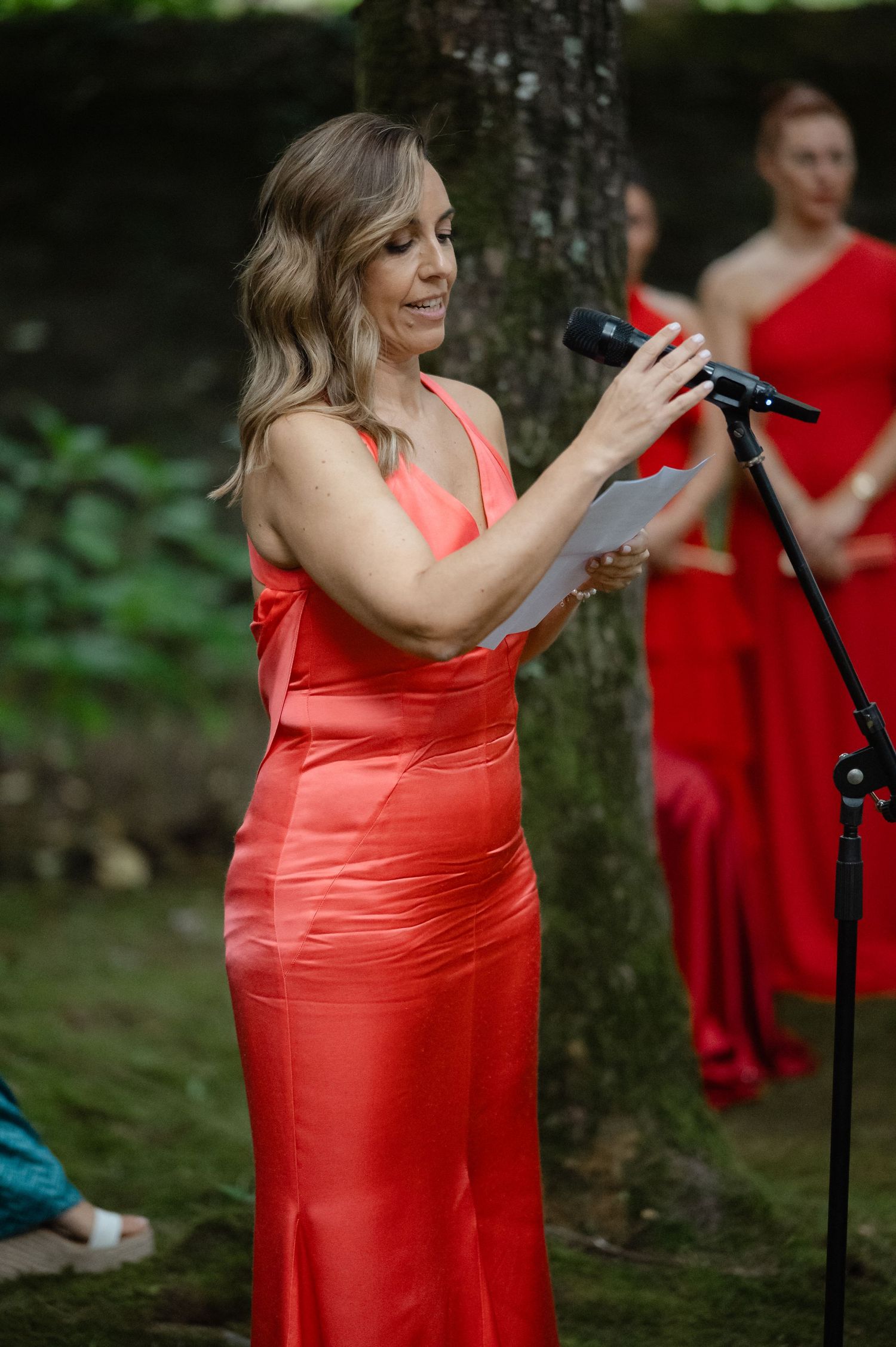 Woman in coral formal dress speaks into microphone at outdoor event with trees in background.