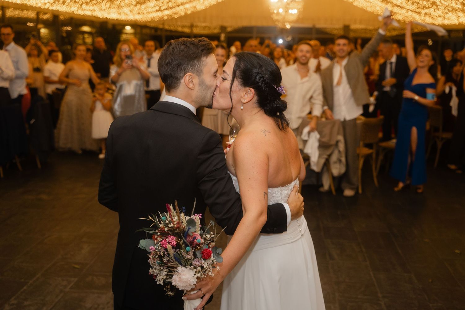 A romantic first dance moment at a wedding reception under twinkling lights with the couple sharing a kiss.