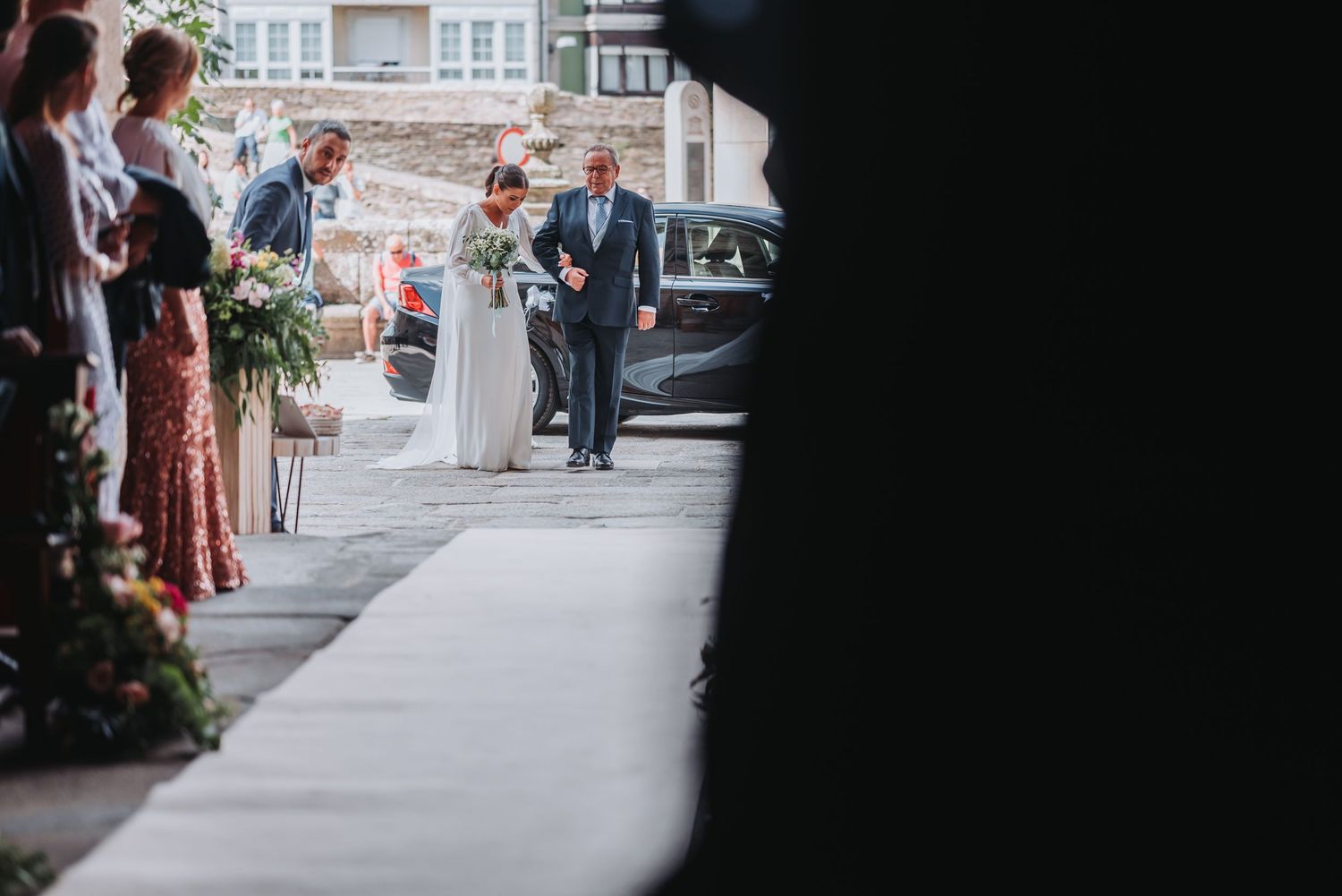 Wedding guests line a sidewalk outside a building as they await the arrival of the wedding party.