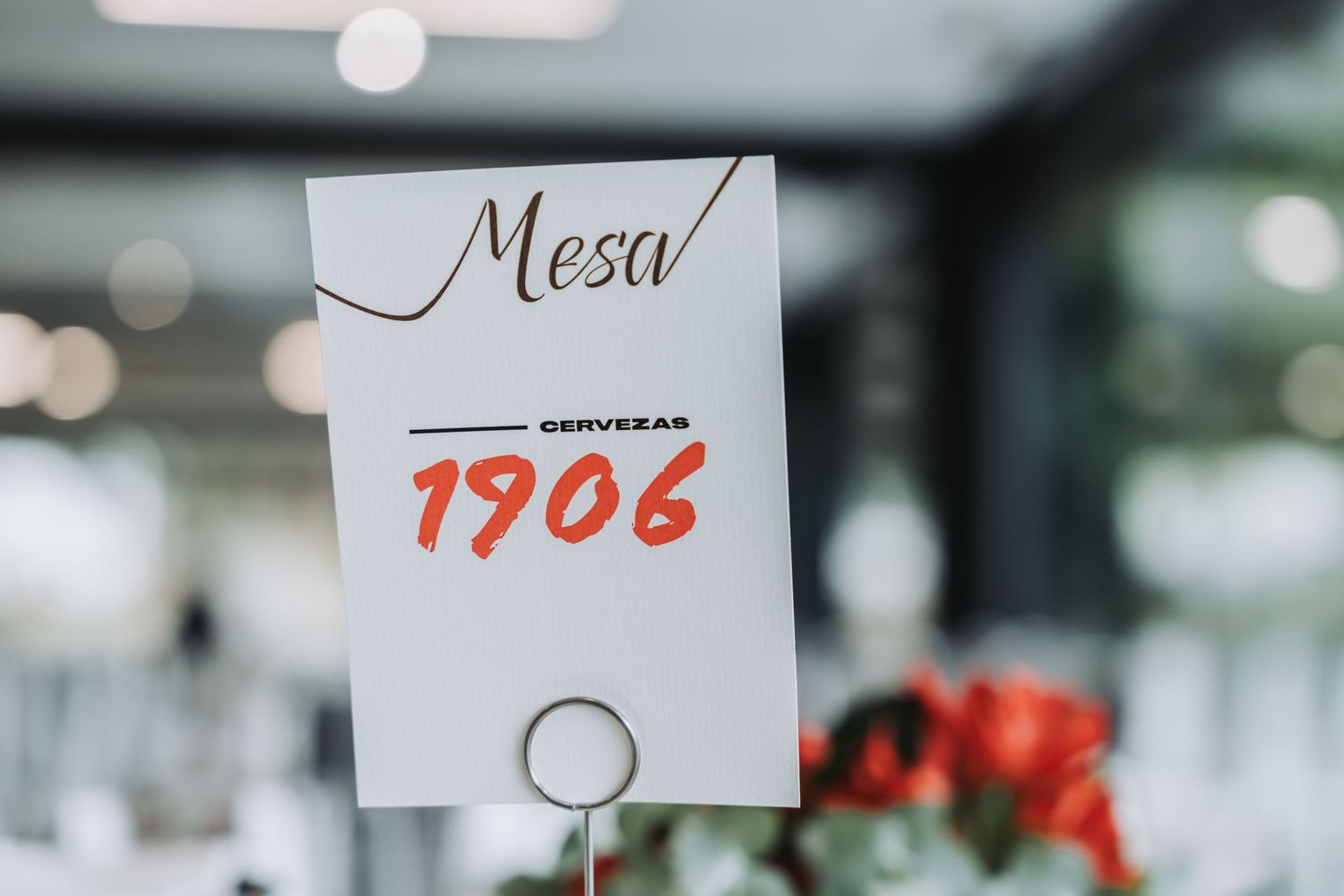 A white table number card displaying 'Mesa 1906' on a metal stand with blurred event decor in the background.