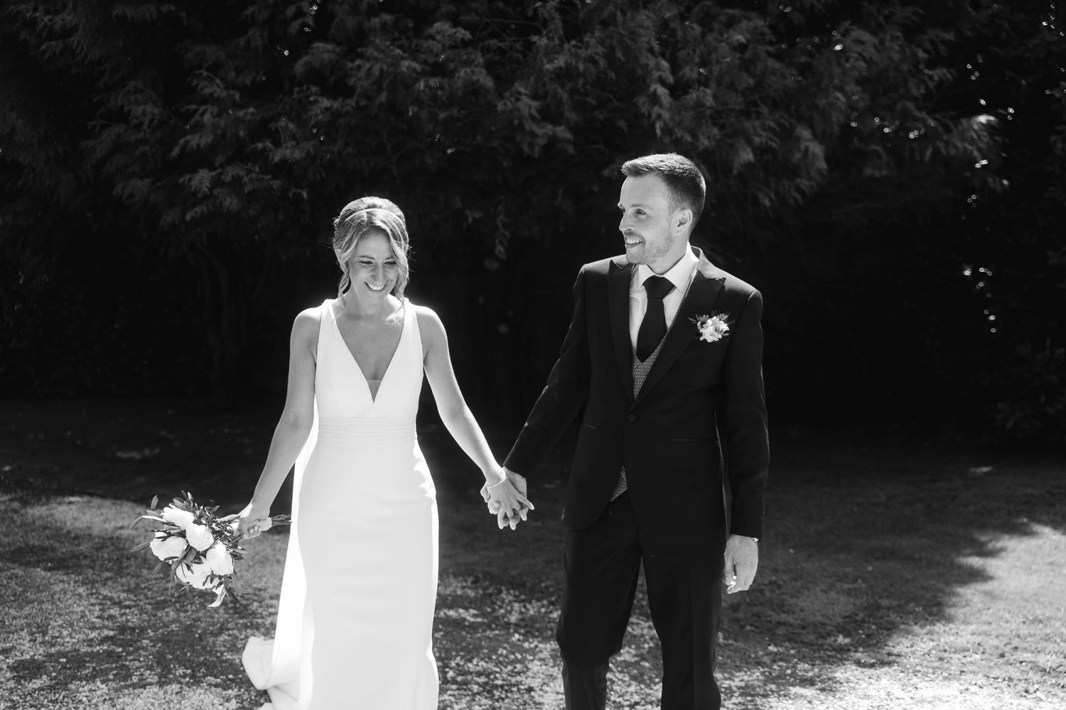 A couple in wedding attire walks hand in hand through a lush outdoor garden setting on their special day.