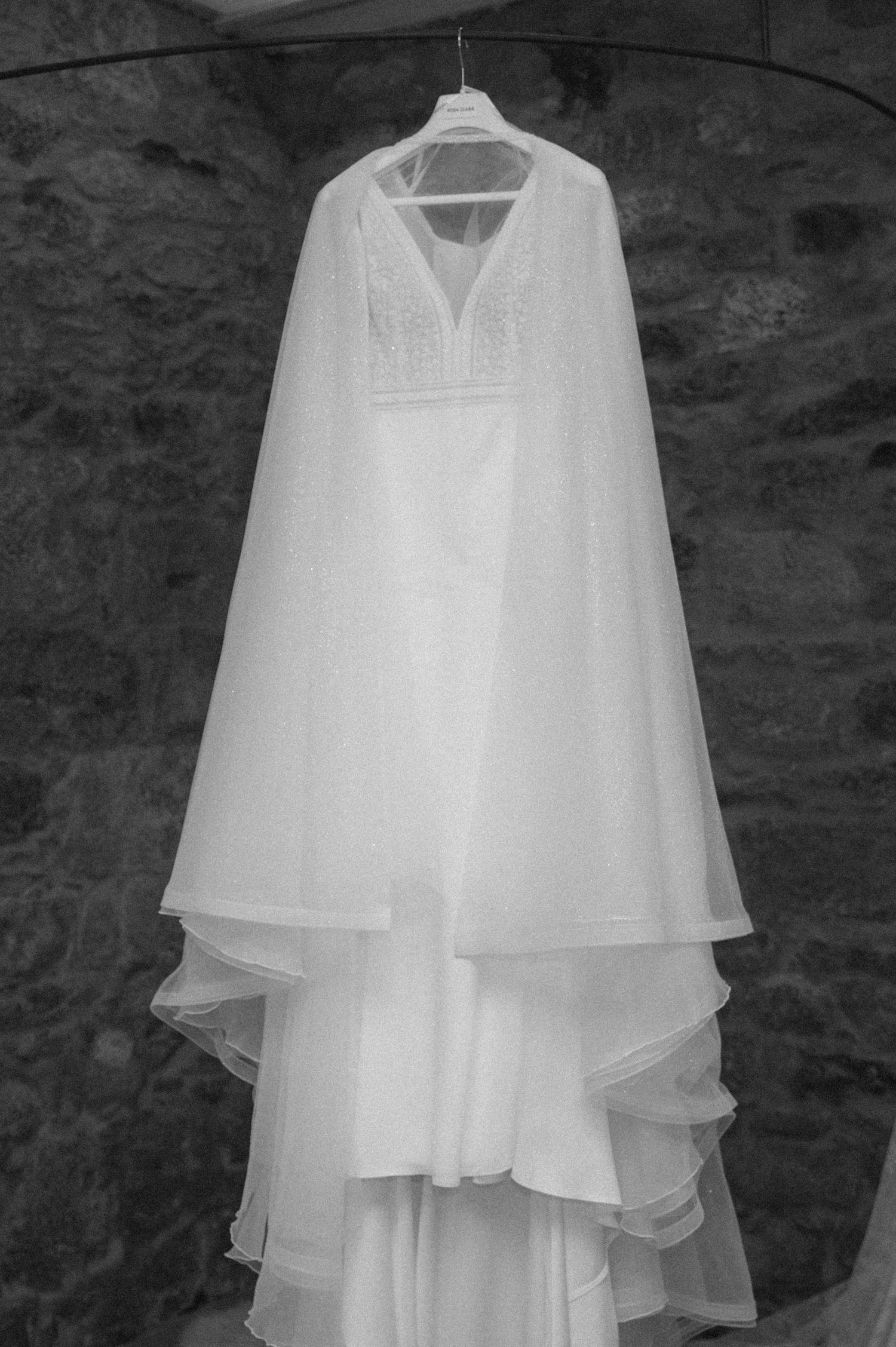 An elegant white wedding dress with delicate details hangs against a stone wall in black and white.