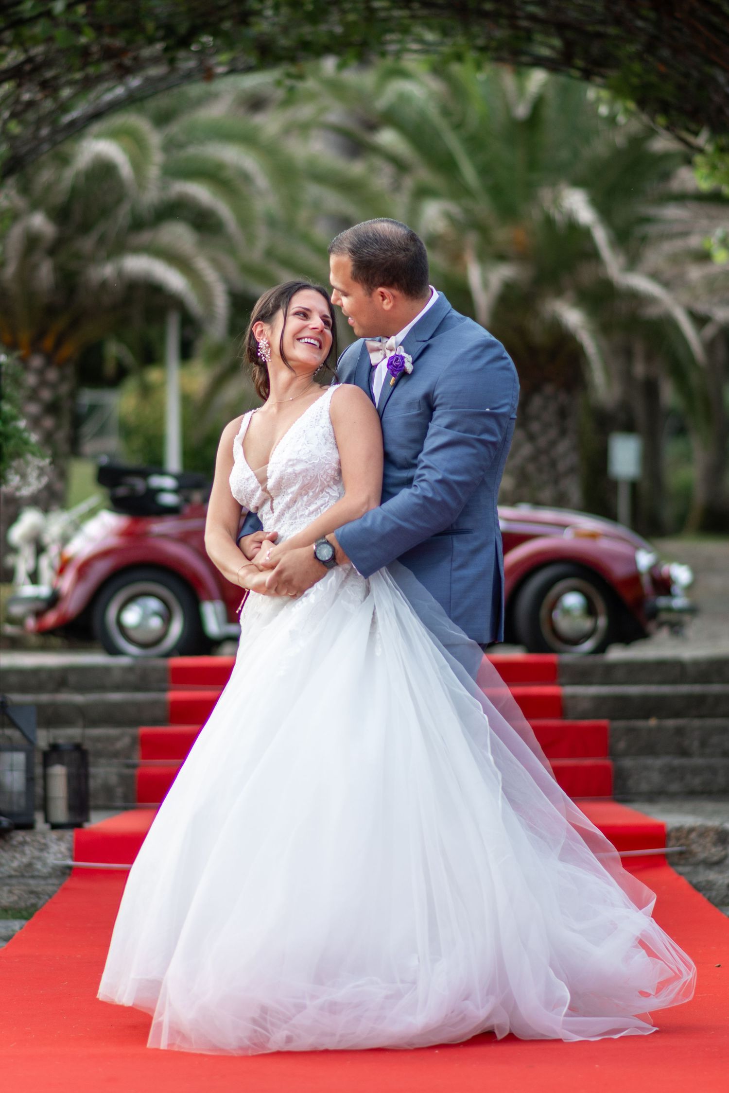A couple in wedding attire poses romantically on red carpet stairs surrounded by palm trees.