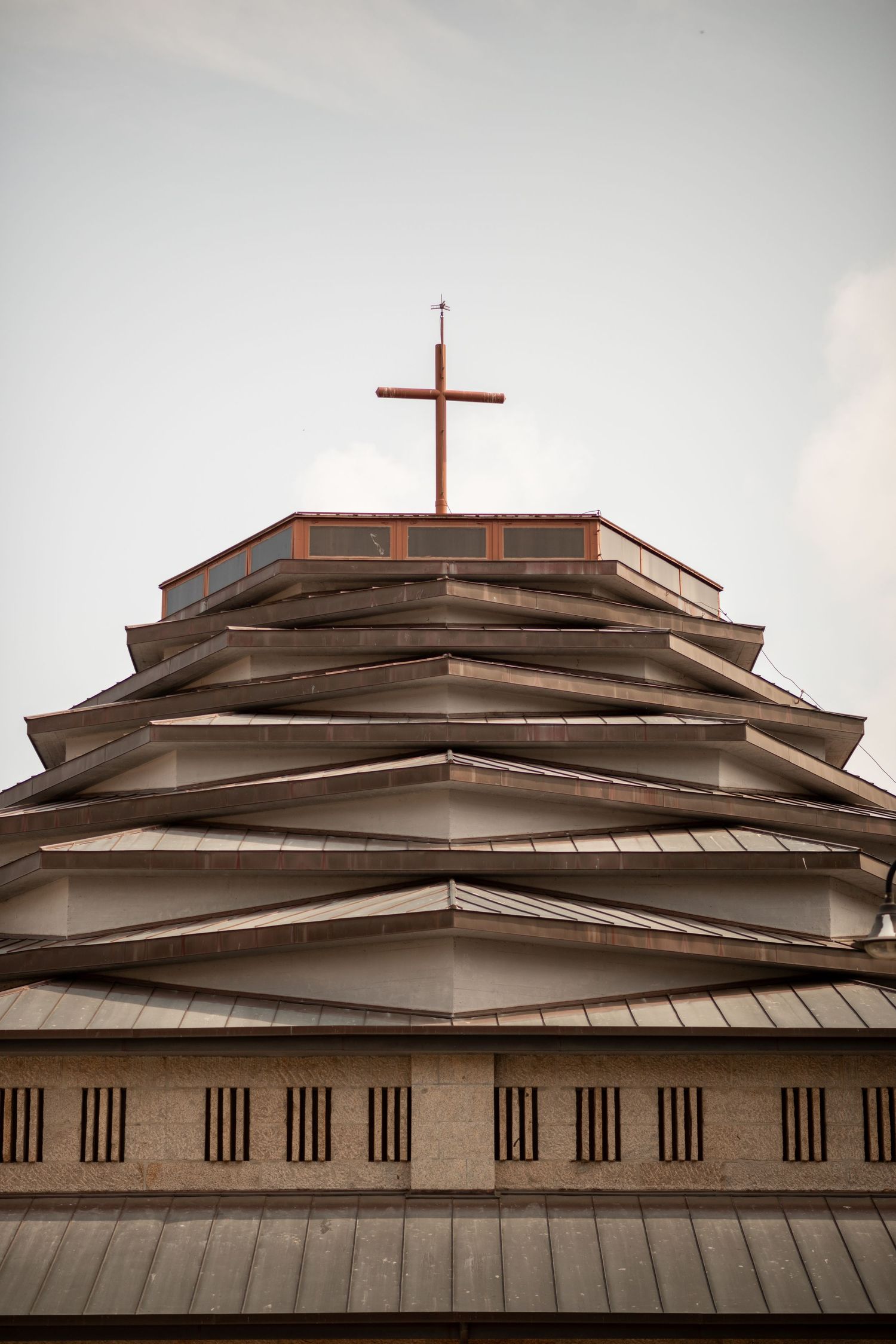 Looking up at a tiered church steeple with wooden cross on top against cloudy sky.