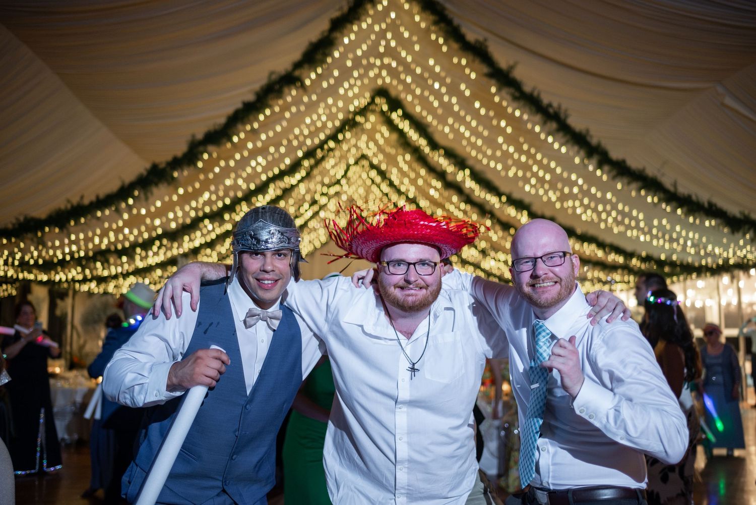 Group of wedding guests dance in a line under twinkling string lights at an indoor reception venue.