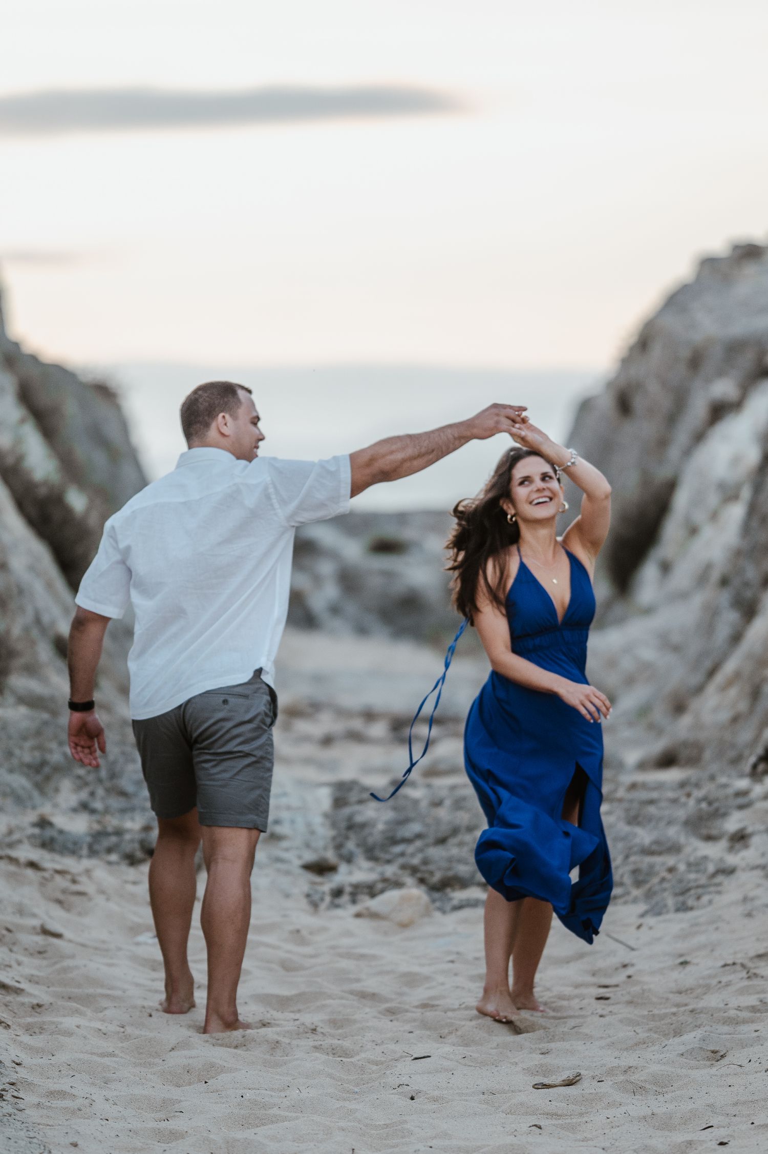 A couple dances together barefoot on a sandy beach at sunset near rocky outcrops.