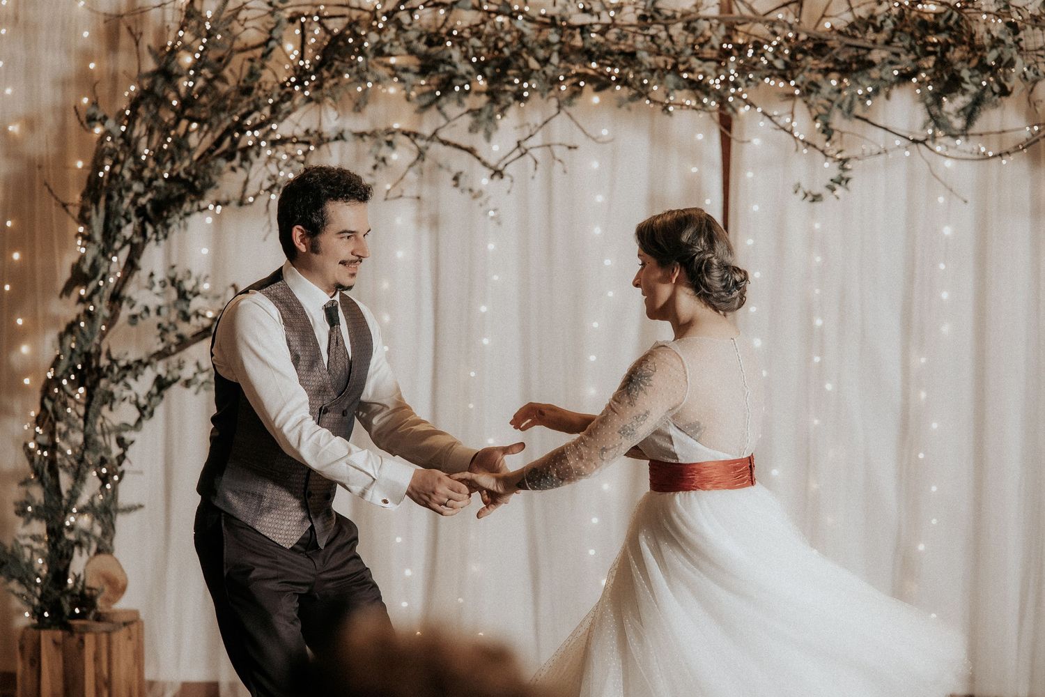 A couple shares their first dance at a wedding reception with twinkling lights and vines decorating the background.