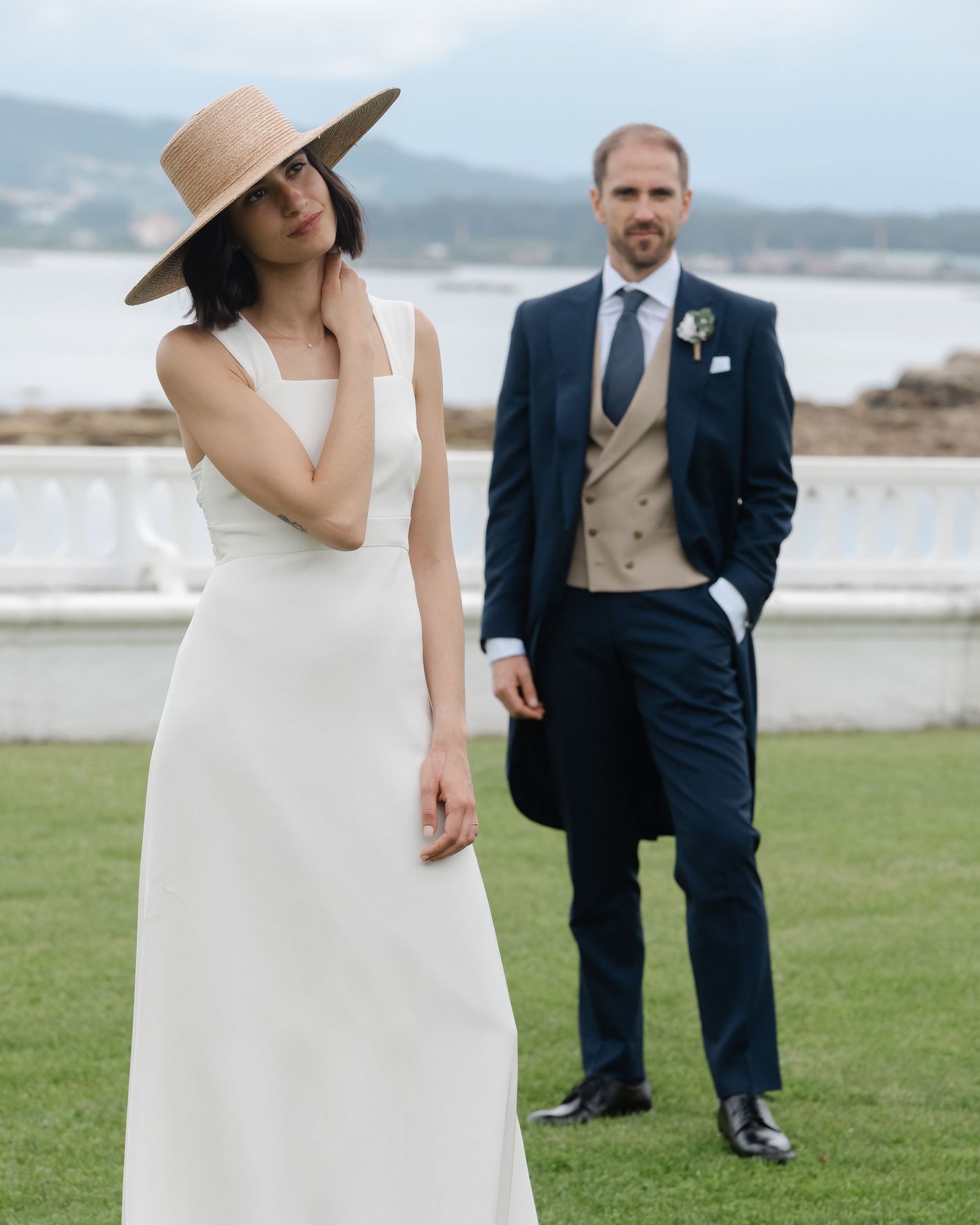A stylish bride in a wide-brimmed hat poses with groom in formal attire by the seaside.Wedding photographer Spain. Galicia.