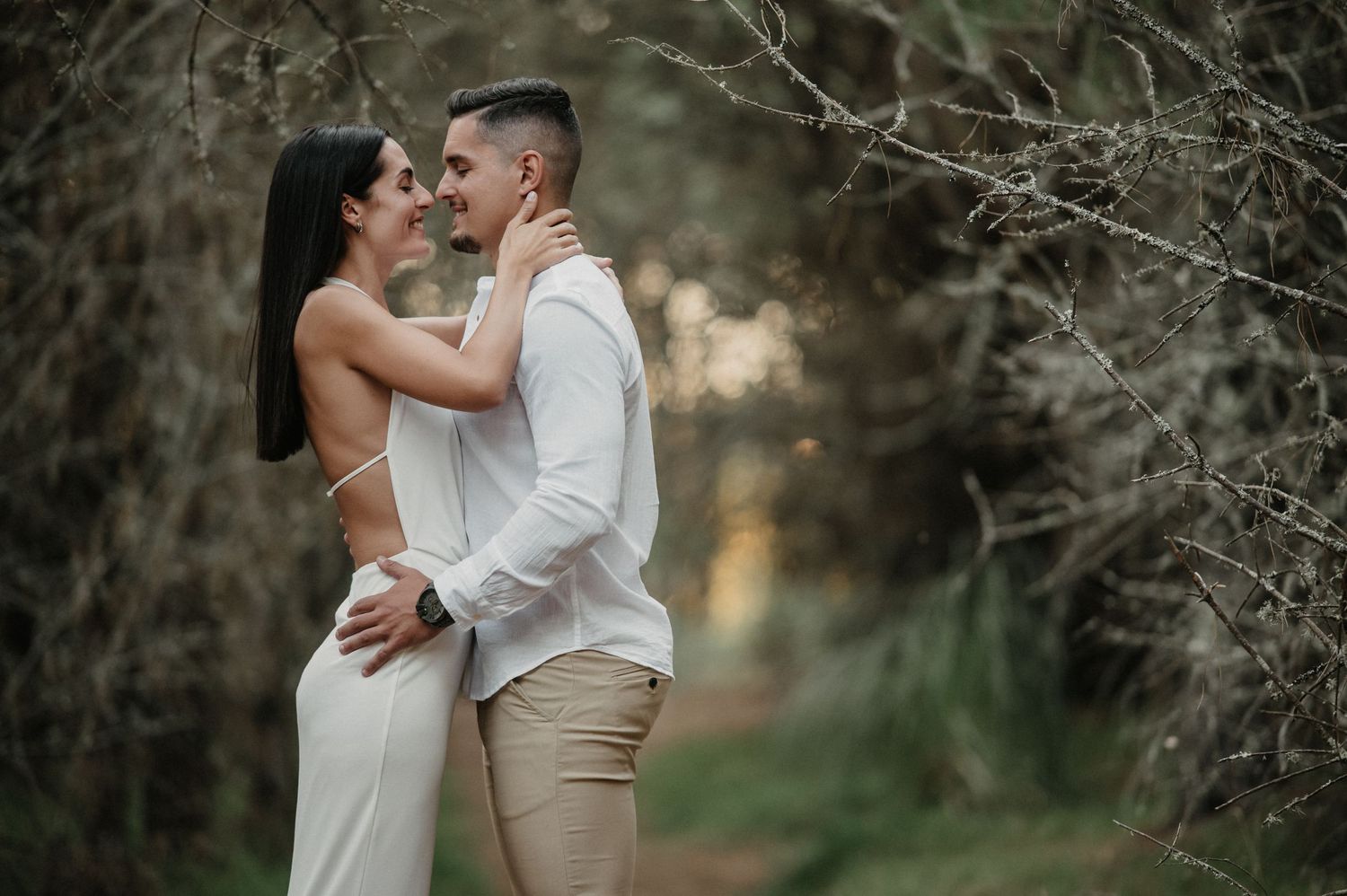 Romantic couple embraces in white attire among moody natural surroundings during outdoor photoshoot at sunset.