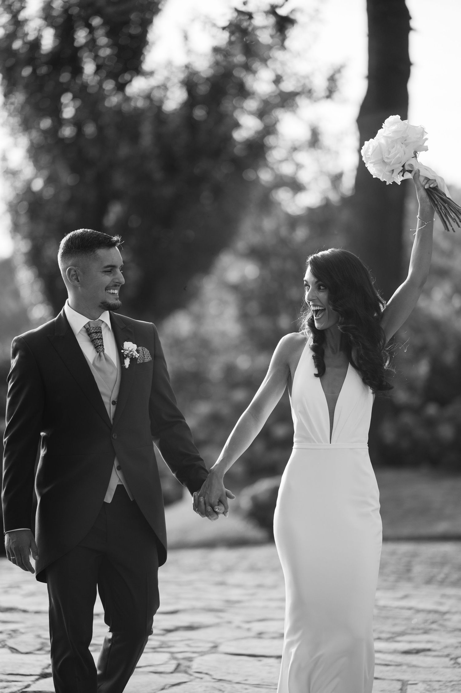 A bride and groom share joyful candid moments walking together in a romantic black and white outdoor setting.