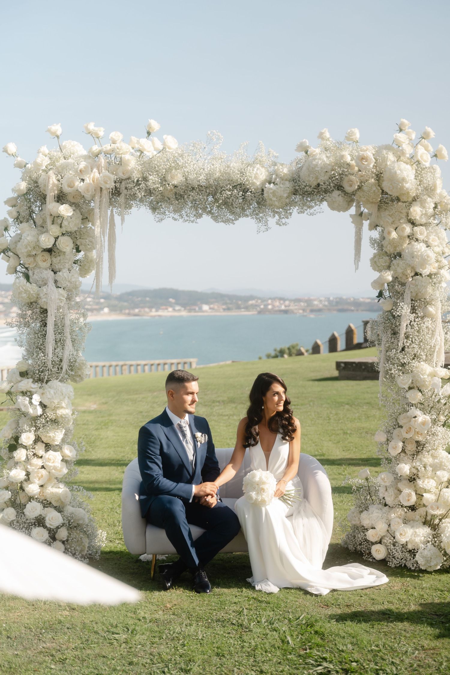 A romantic wedding couple sits on grass beneath elegant white floral arches with an ocean view in the background.