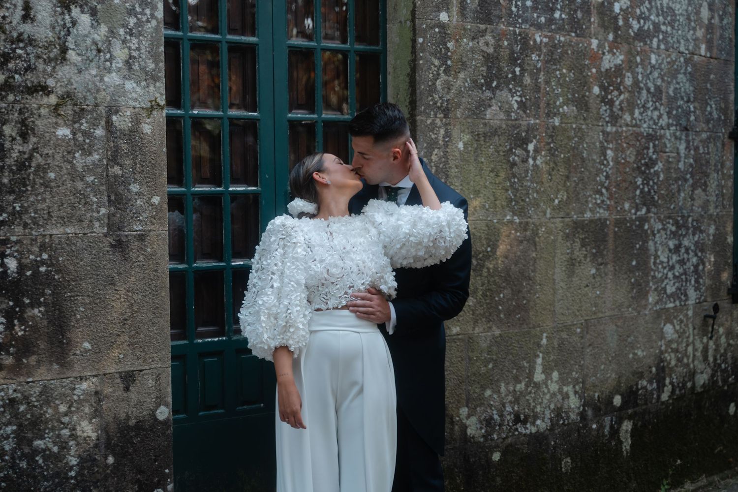 A couple in white wedding attire embrace and dance together against a rustic stone wall with barred windows.