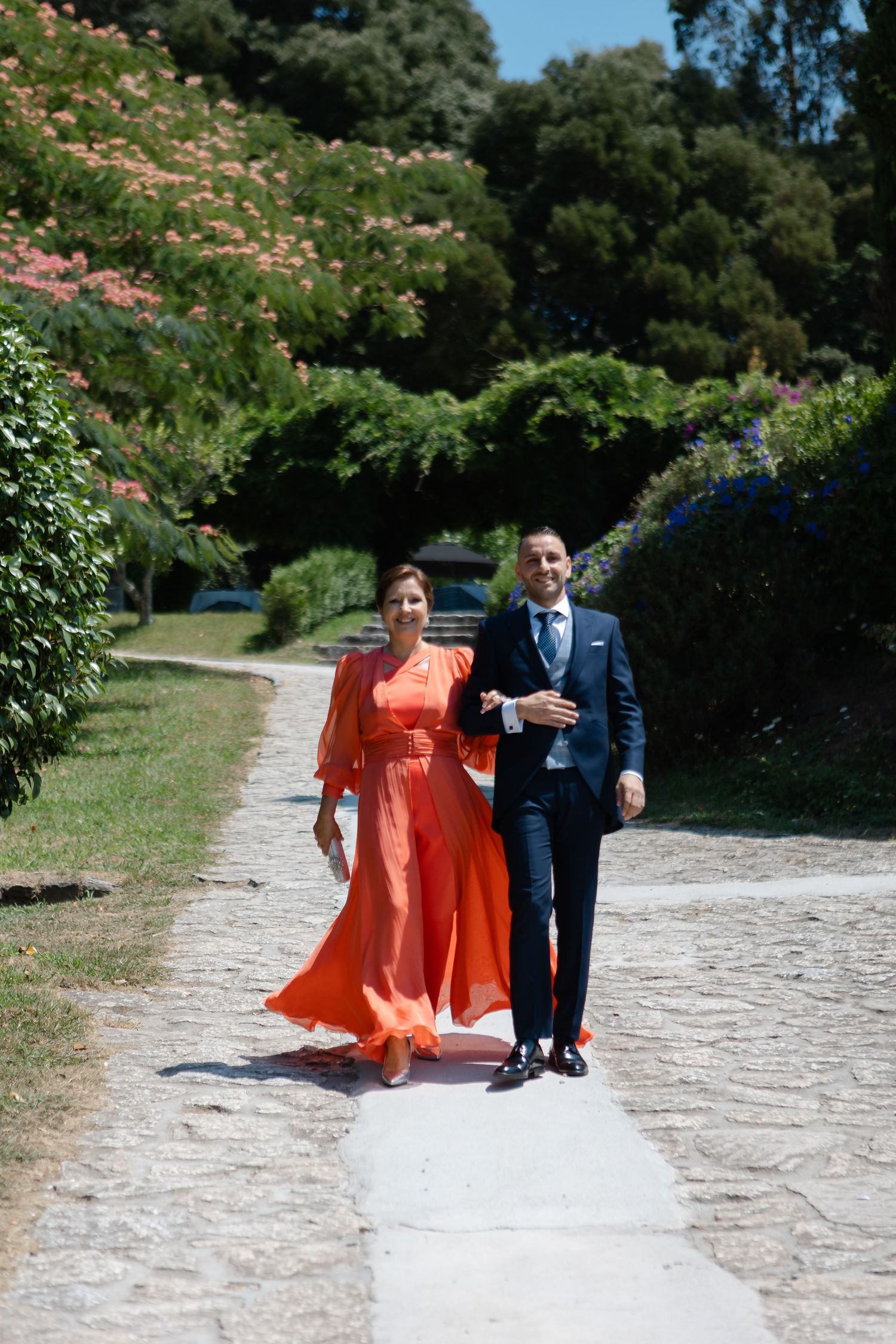 A couple walks along a scenic stone path with the person in orange wearing a flowing dress that catches the breeze.