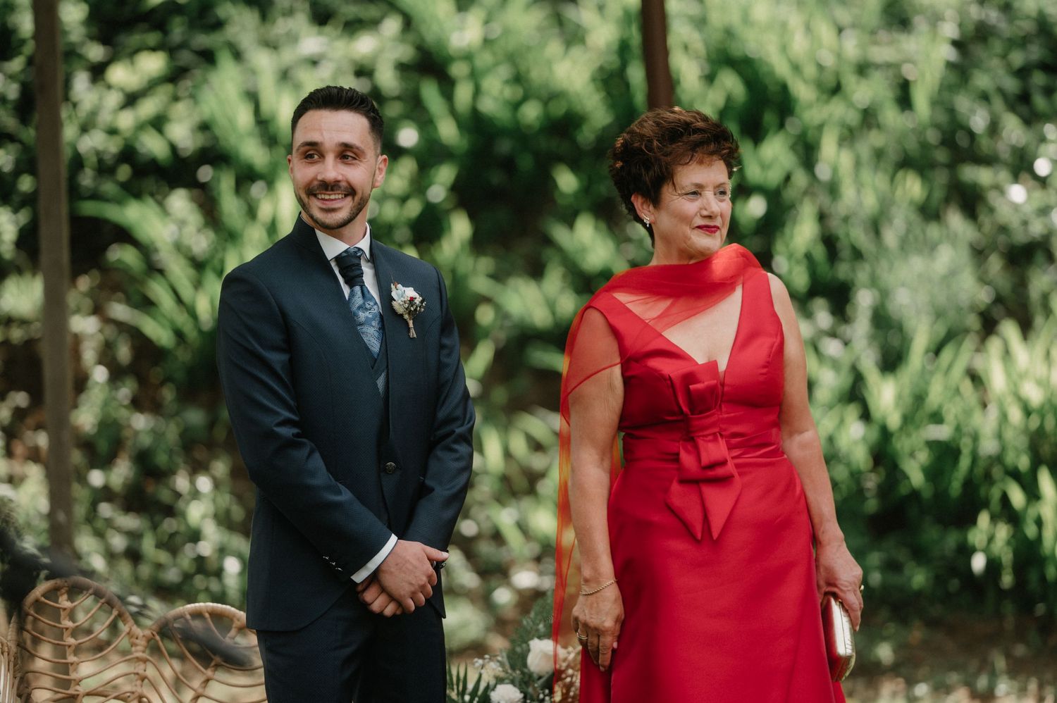 Wedding ceremony attendees in formal attire gathered outdoors with greenery in the background.