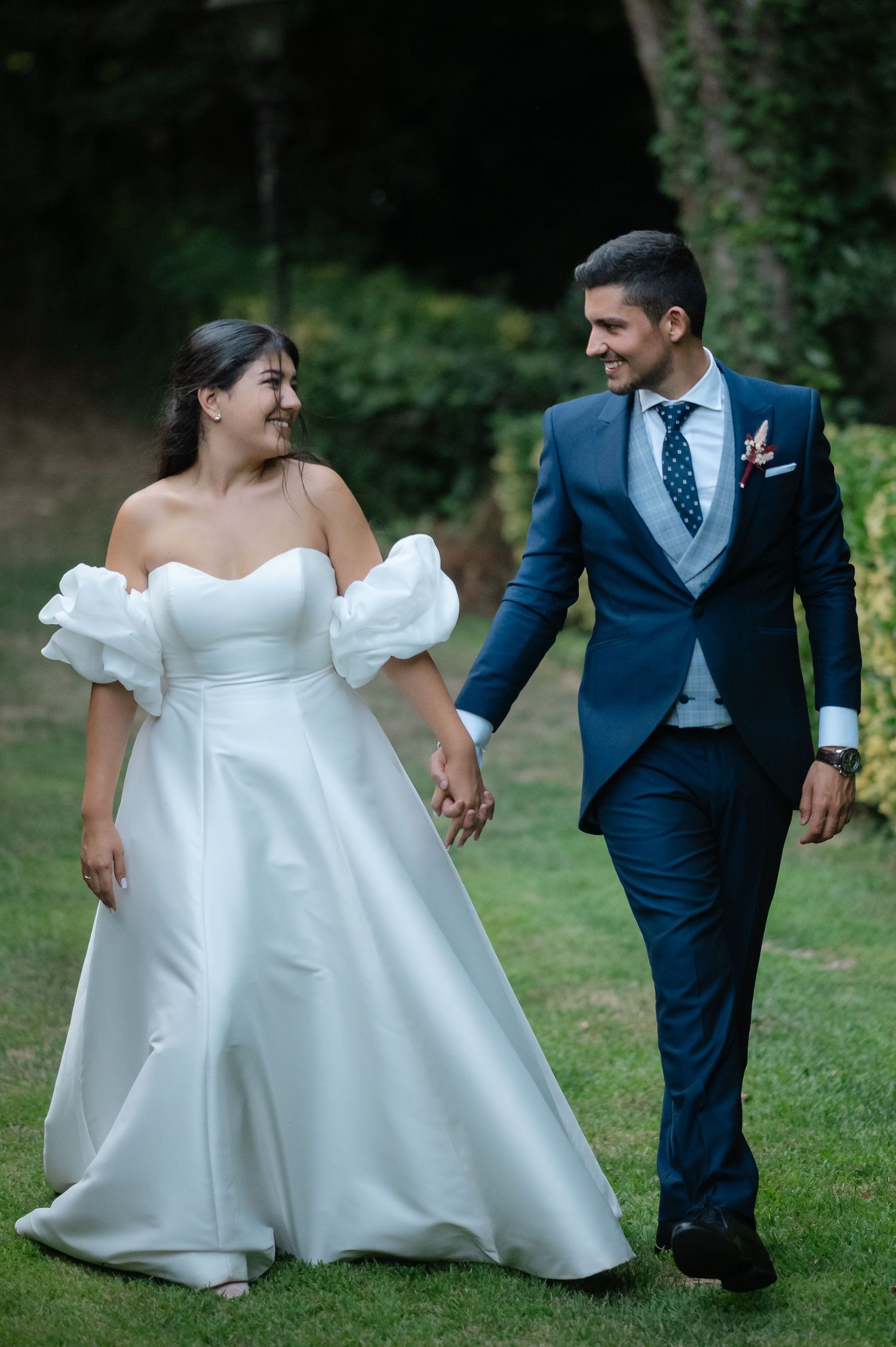 A couple in wedding attire walk and laugh together on a grassy lawn, with the bride in a flowing white gown.
