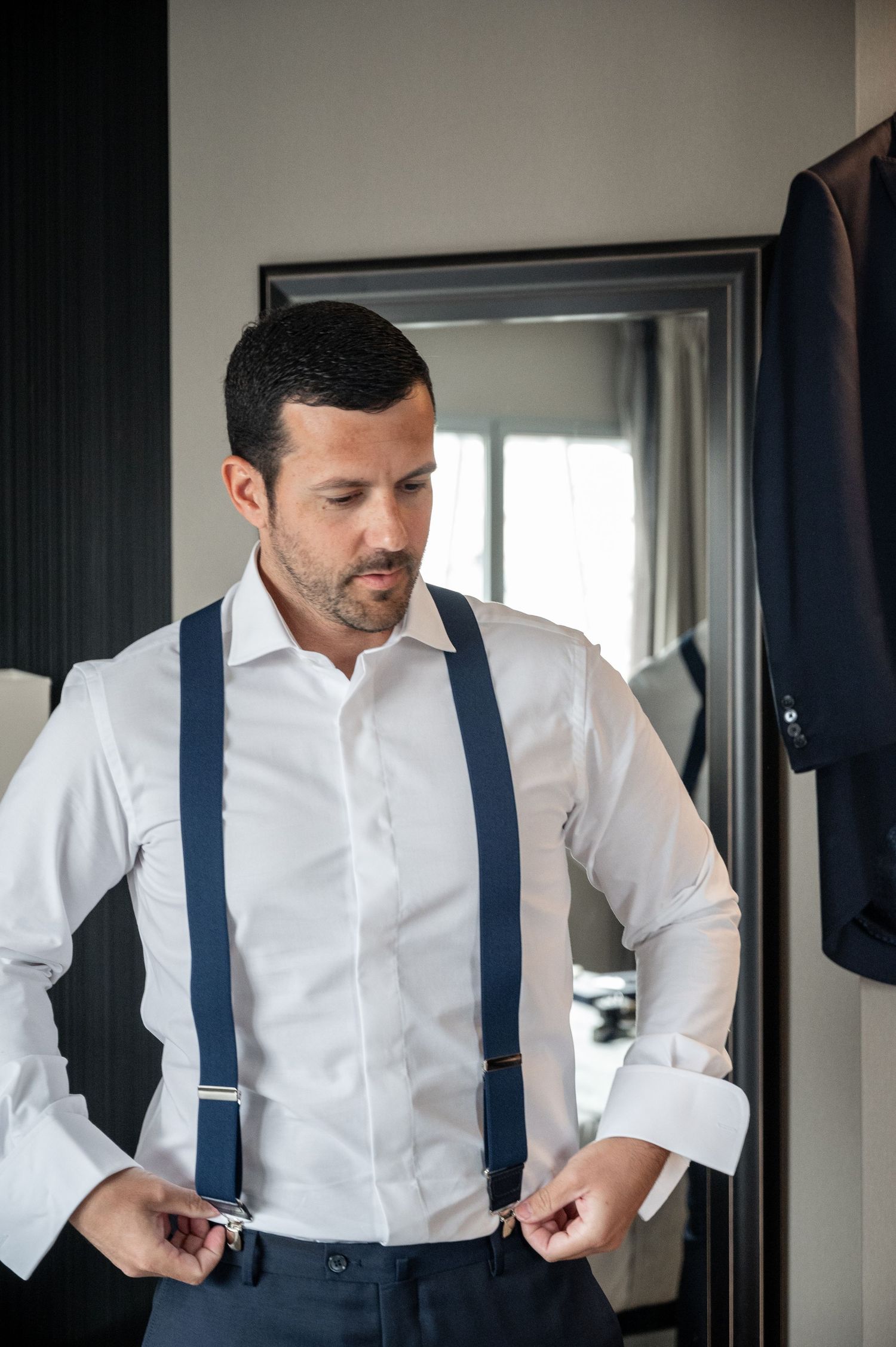 A man in formal attire adjusts his navy blue suspenders while wearing a crisp white dress shirt.
