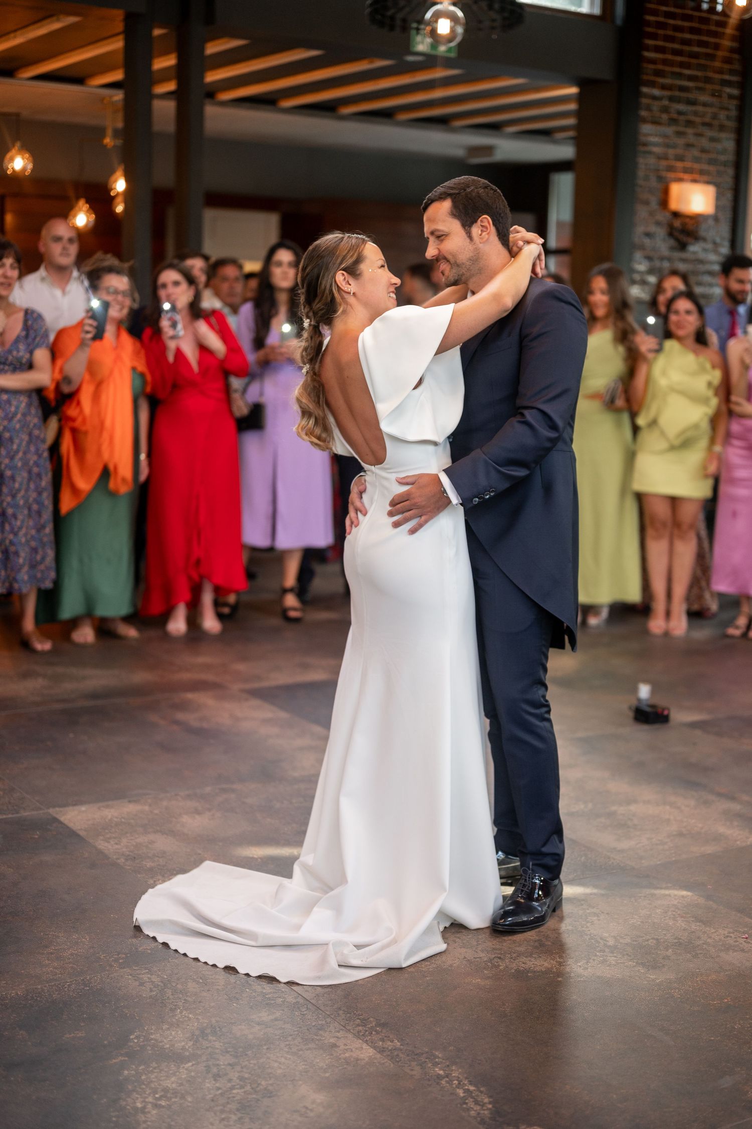 A couple shares their first dance at a wedding reception while guests in colorful dresses look on from the background.