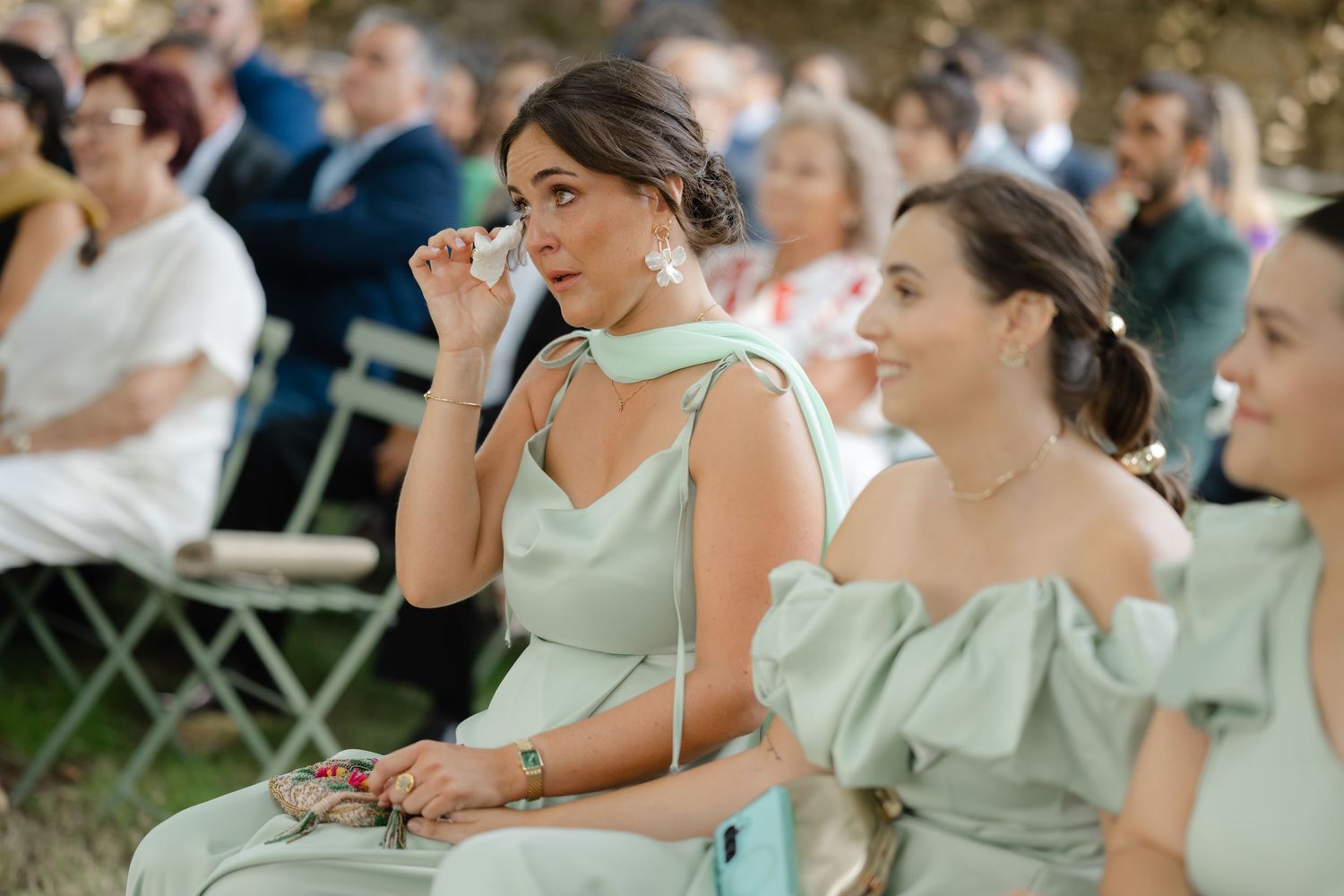 Emotional guests in mint green bridesmaid dresses watch an outdoor wedding ceremony from folding chairs.