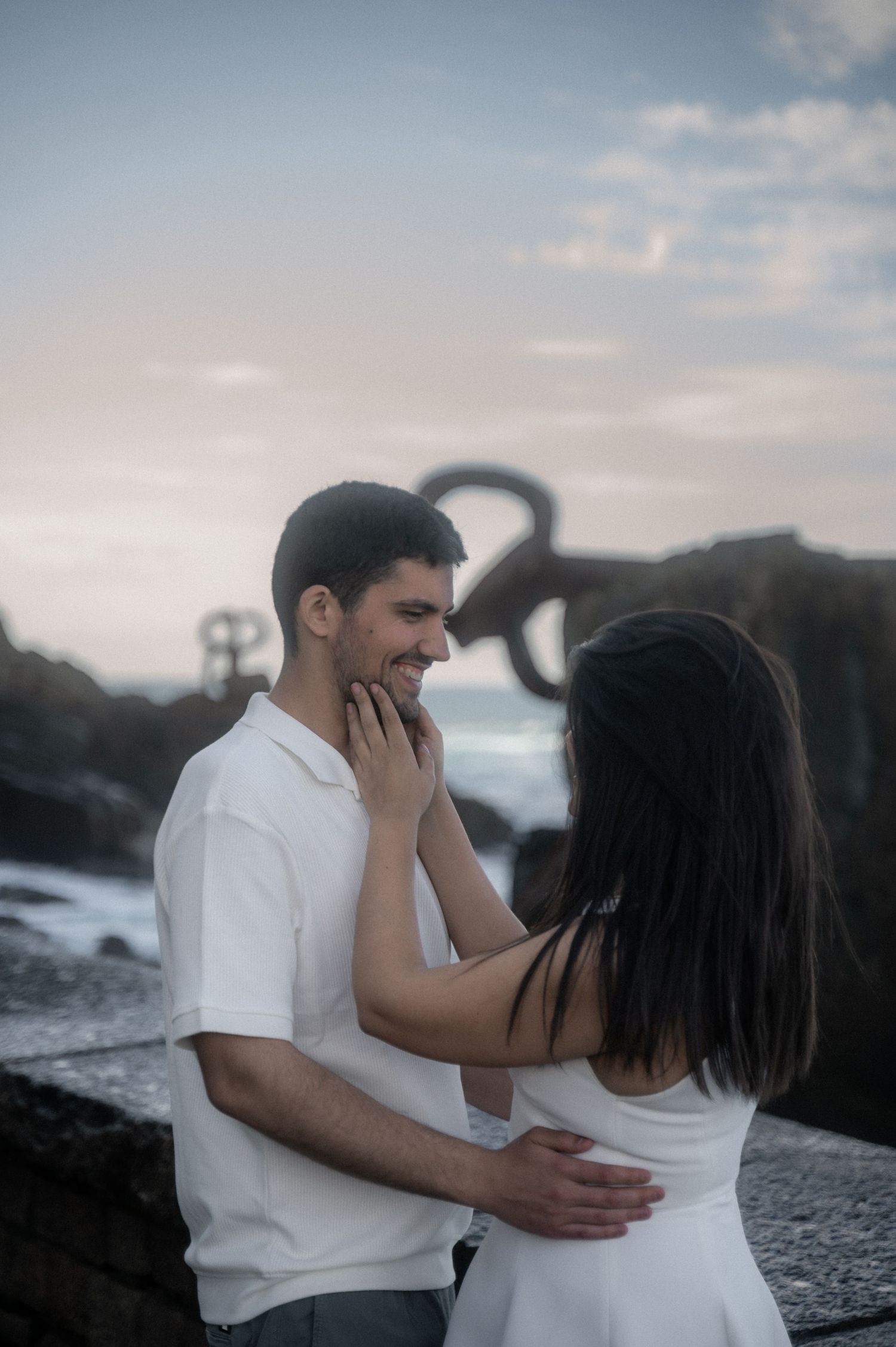 Romantic couple in white outfits embrace against a moody coastal backdrop at sunset.