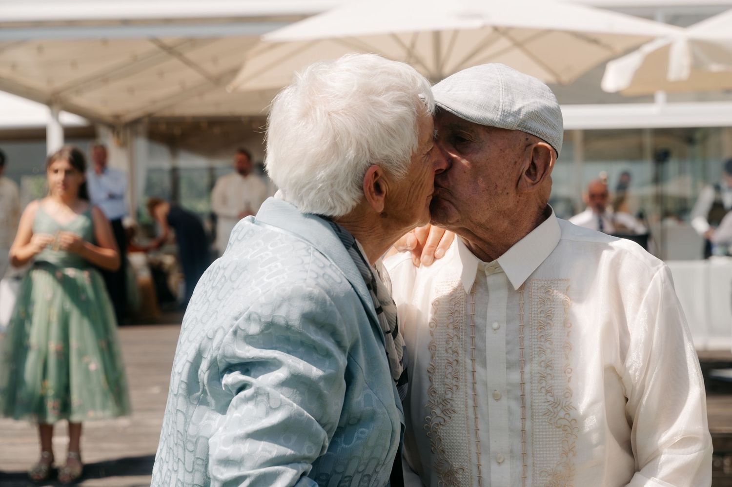 An elderly couple in light colored clothing shares a tender moment while sitting together outdoors.