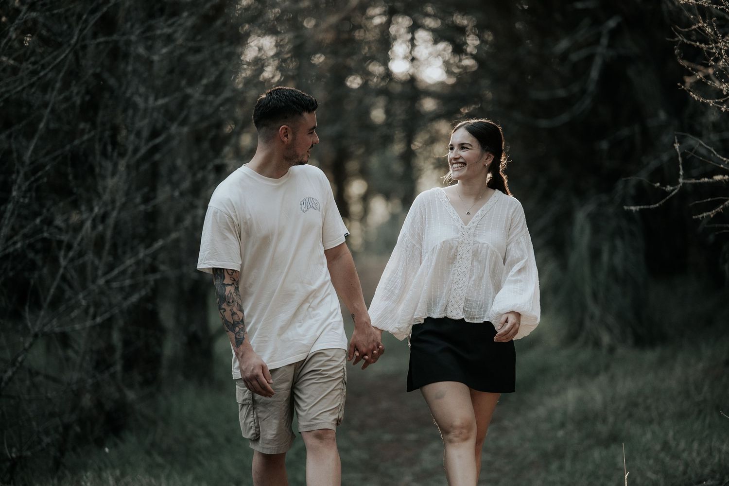 A couple walks hand in hand through a moody forest setting during golden hour.