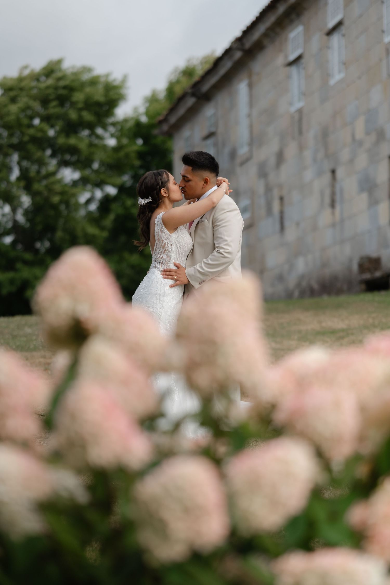 Romantic wedding couple embraces behind soft pink flowers in foreground against stone building exterior.